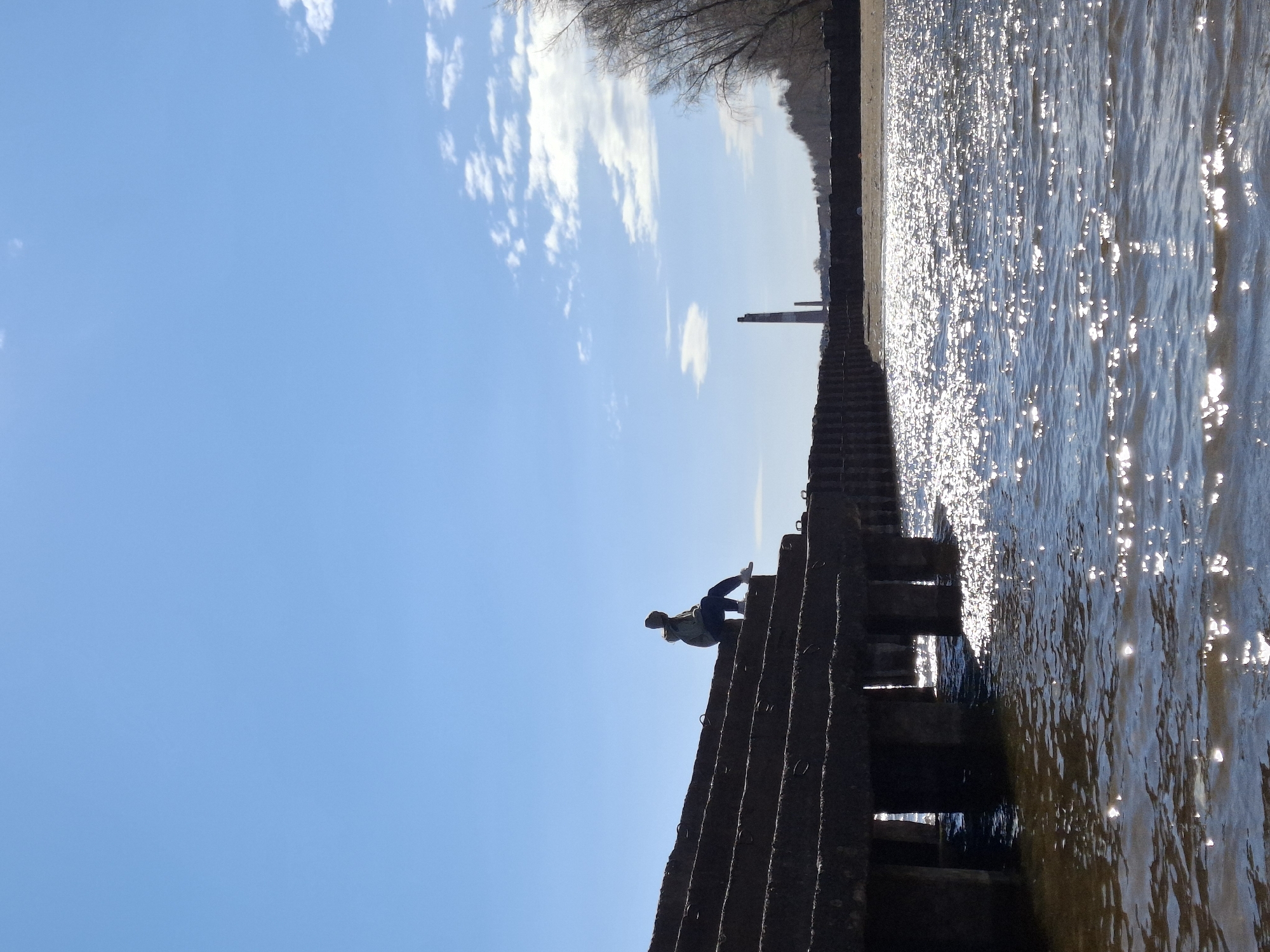 A person is perched on the edge of a concrete structure, possibly a pier or embankment, in what appears to be the city of Chaikovsky, Russia. The structure has a series of step-like indentations and is partly submerged in water. The water's surface reflects sunlight, creating a glittering effect. Beyond the concrete structure is a line of trees or vegetation, suggesting a shoreline. The sky is blue with scattered clouds. The person is facing away from the viewer and their posture suggests they may be looking at the water or the horizon. The image is oriented so that the sky is on the left and the water on the right.