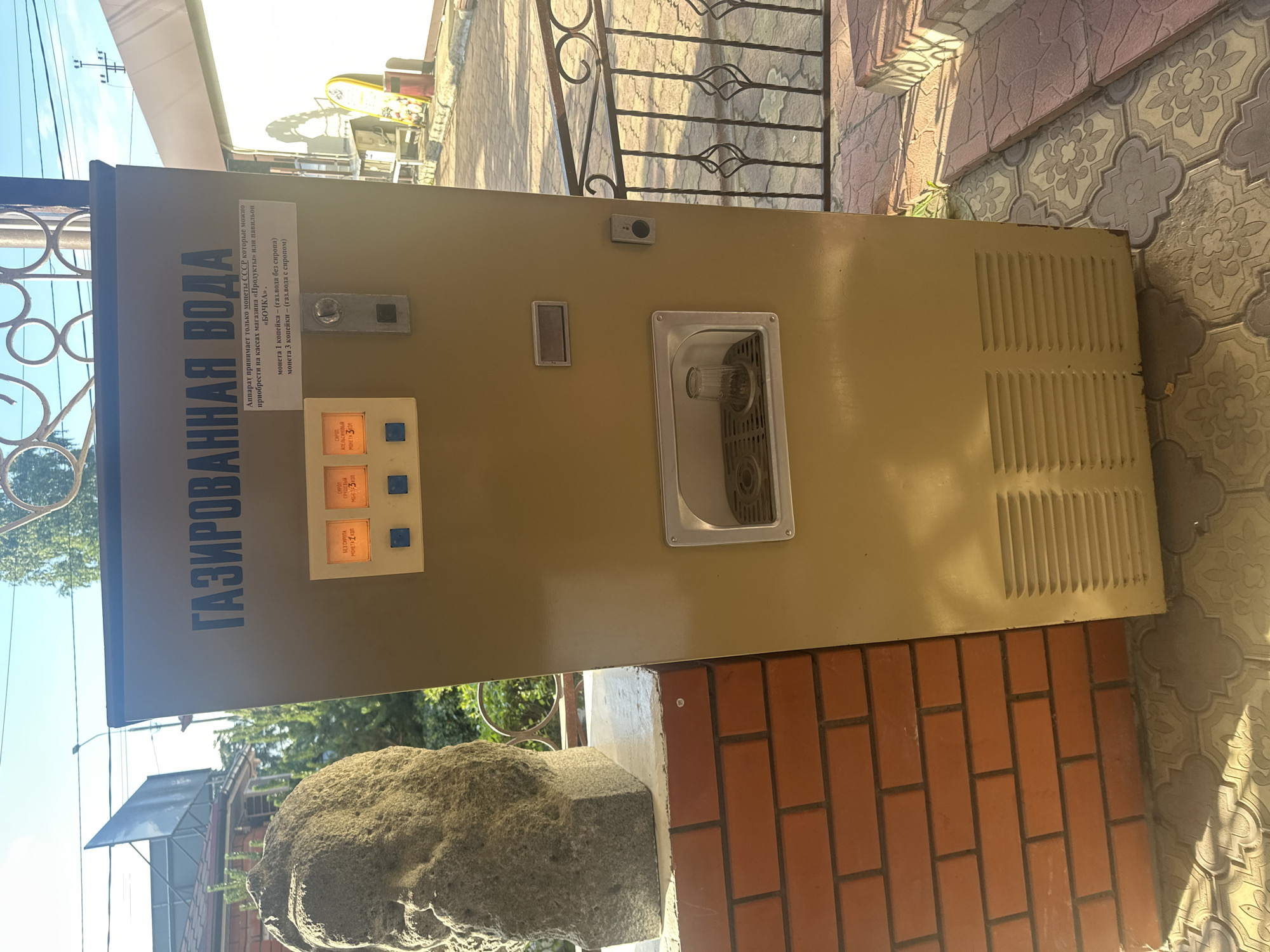 This is a medium-shot, eye-level, outdoor photograph of a vintage Soviet-era carbonated water dispenser in Kablukovo, Russia. The dispenser is a tall, beige, metal cabinet with Cyrillic text across the top that reads "ГАЗИРОВАННАЯ ВОДА" (Carbonated Water). Below the text, a smaller white label indicates that the machine accepts USSR coins and specifies prices for plain and flavored water. To the left of the label are two illuminated orange buttons, each labeled with a coin denomination and description: "БЕЗ СИРОПА МОНЕТА 1 КОПЕЙКА" (Without Syrup, 1 Kopeck Coin) and "С СИРОПОМ АПЕЛЬСИНОВЫЙ МОНЕТА 3 КОПЕЙКИ" (With Syrup, Orange, 3 Kopeck Coin). There is a coin slot in the center of the machine, and a glass is present in the dispenser area.
To the left of the dispenser, a rough, stone bust of a person's head and shoulders stands on a brick wall. In the background, a metal fence with ornate scrollwork is visible, with patterned paving stones on the ground. The scene appears to be in daylight, with shadows cast by the sun. The overall impression is one of a public space, likely an outdoor waiting area or a pathway.