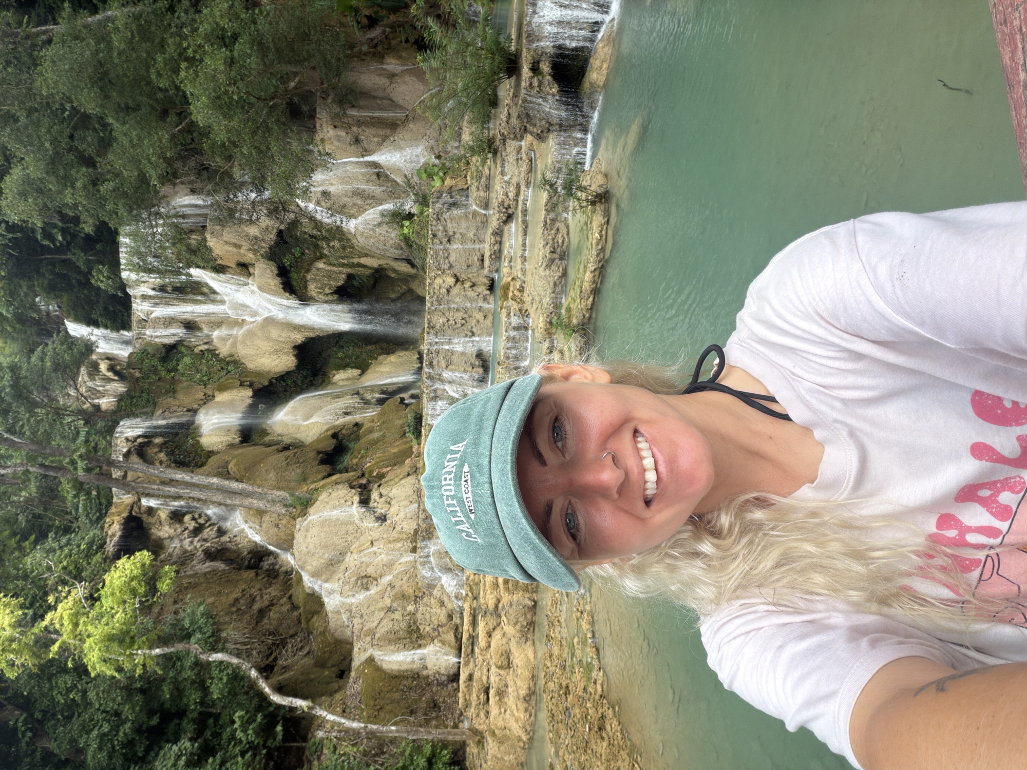 A woman with blonde hair, wearing a light pink t-shirt and a green baseball cap, smiles at the camera in a selfie. She is positioned in the foreground, with a multi-tiered waterfall and turquoise water in the background. The waterfall cascades over limestone formations, surrounded by lush green foliage and trees. The scene appears to be at Kuang Si Falls, a popular tourist attraction located about 29 kilometers (18 miles) south of Luang Prabang, Laos, and is not located in the city of Thapene. The woman has a nose ring and visible tattoos on her arm. The water is clear enough to see the bottom in some areas.
