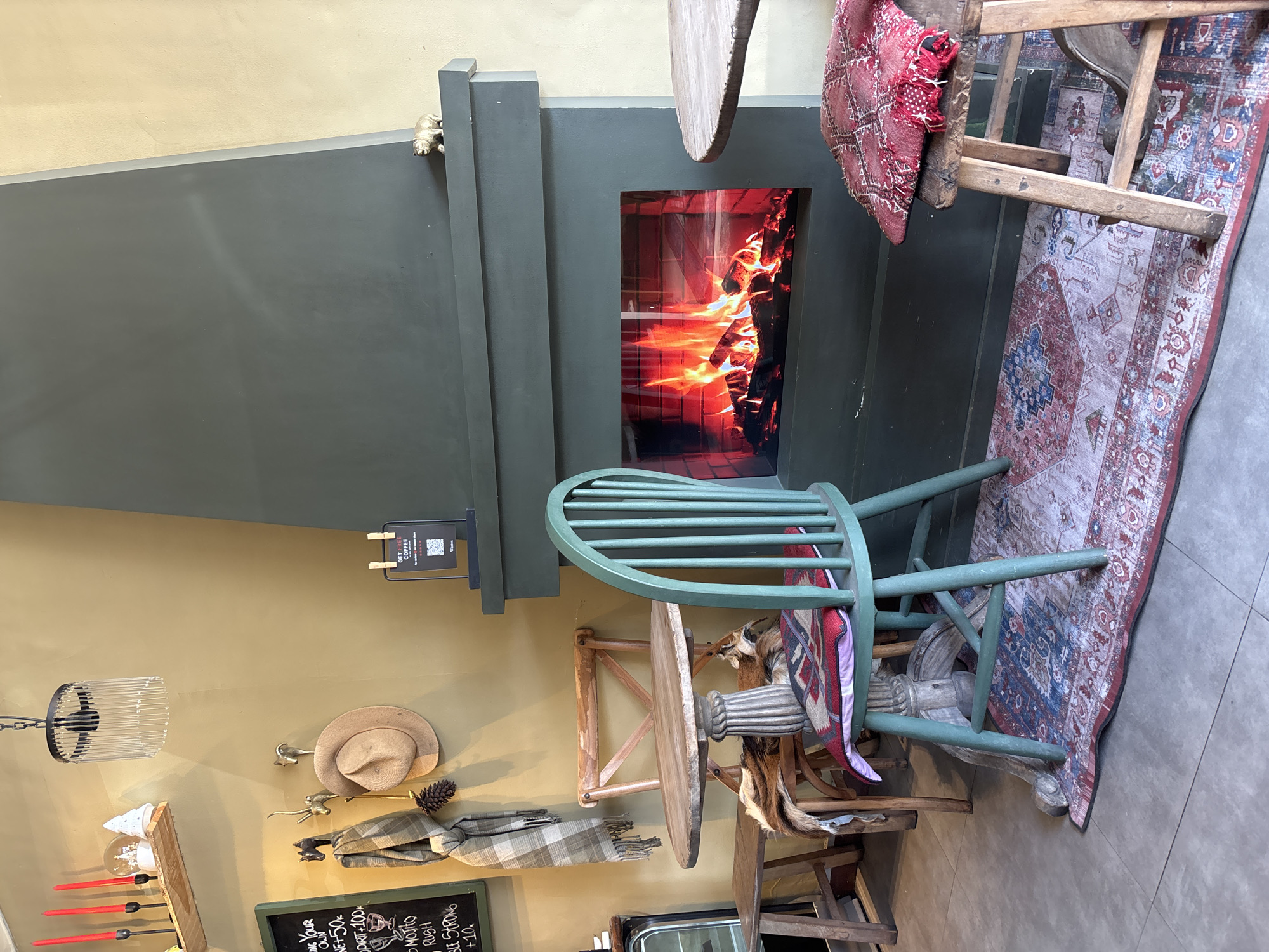 The image depicts an interior scene, possibly in a café or restaurant in Kuta Selatan, Indonesia. The lighting is diffused and warm. A green fireplace is visible, displaying a simulated fire. Two wooden chairs are present; one is green, and the other has a red patterned cushion. A small round table is positioned beside the fireplace. The floor is covered by a patterned rug. On the wall, there are various decorative items, including a hat, a scarf, and a blackboard with writing in Indonesian, advertising drinks such as "MOJITO." There's also a sign that says "GET FREE COFFEE."
