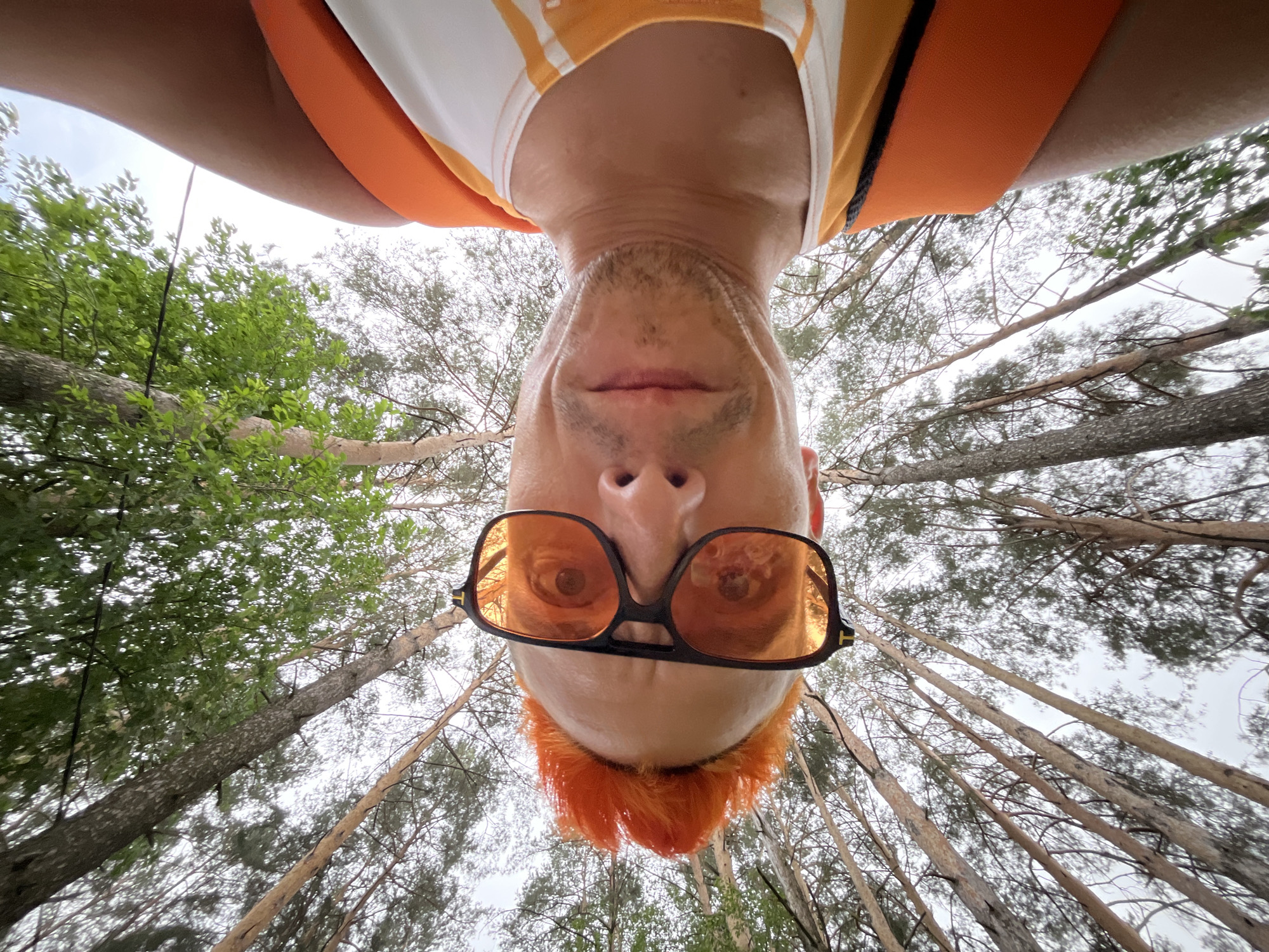 A man wearing orange-tinted glasses and an orange and white tank top is pictured in an upside-down perspective. He has short orange hair styled upwards. The background consists of tall trees stretching upwards, with green foliage on the left side and bare branches on the right, suggesting a forest setting. The image was taken outdoors in daylight. This image was most likely taken somewhere in the city of Chaikovsky, Russia.