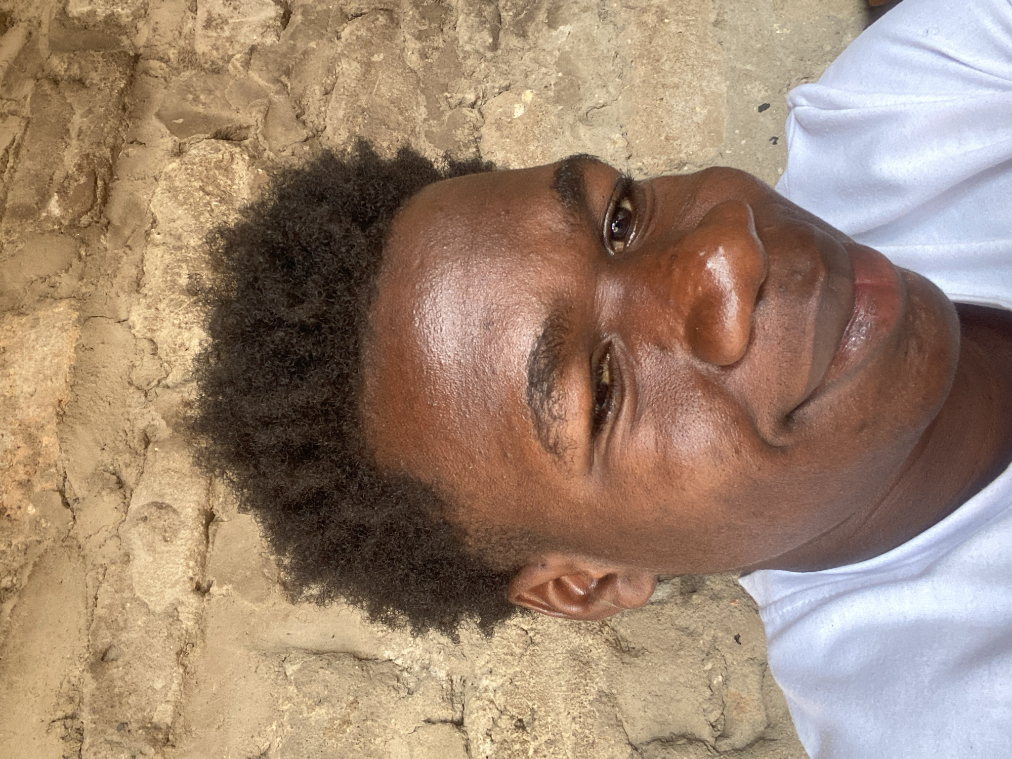 An individual with dark skin and short, dark, curly hair is lying on a textured, possibly concrete or stone surface, wearing a white t-shirt. The person's face is turned towards the camera, and their expression appears relaxed. The background is a rough, uneven surface. The image might have been taken in Mararaba, Nigeria, though there are no specific identifiers visible.