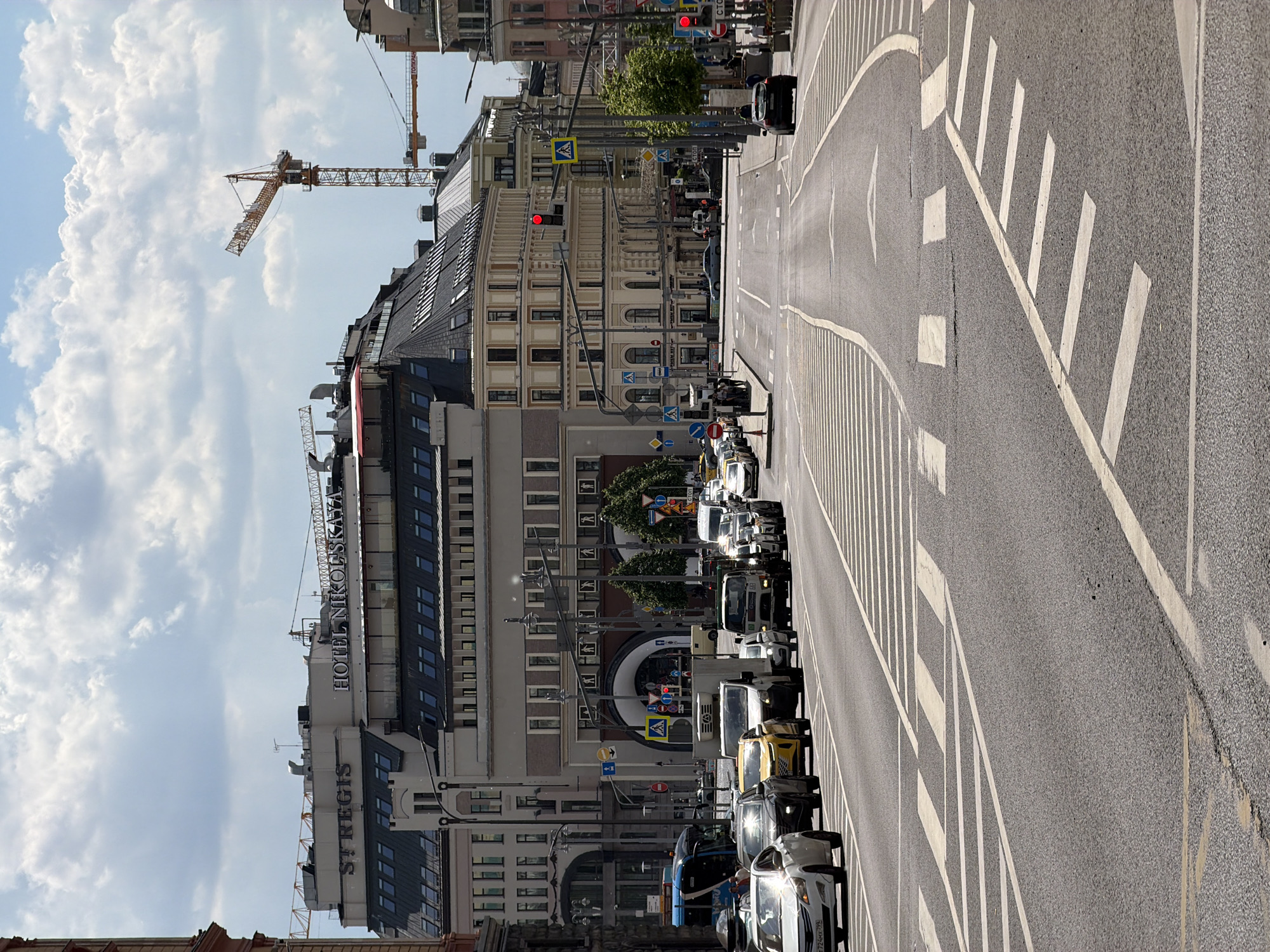 This image, set in Moscow, Russia, captures a broad city street with significant urban architecture and active traffic under a partly cloudy sky. The perspective is slightly elevated, looking down a wide, multi-lane asphalt road that curves gently to the right in the distance.
On the left side of the street, a large, prominent building complex stands, identified by the visible text "ST REGIS" and "HOTEL NIKOLSKAYA" on its facade, indicating it is the St. Regis Moscow Nikolskaya hotel. This building features a dark, sloped roof and multiple windows. Beyond it, several older, ornate buildings with lighter colored facades are visible, characteristic of Moscow's architecture. Above these buildings, two large yellow construction cranes extend into the sky, suggesting ongoing development or maintenance in the area. One crane bears Cyrillic text, likely "Галичанин," identifying its manufacturer.
The road itself is clearly marked with white lines, including solid and dashed lane dividers, turning arrows, and a distant pedestrian crossing (zebra stripes). Numerous vehicles populate the street; some are parked along the left curb, while others form a line of traffic further down, with a red traffic light visible. The traffic appears moderate. Various street signs, some with Cyrillic text (e.g., pedestrian crossing, yield signs), are mounted on poles along the roadside. A few green trees provide touches of greenery amidst the urban landscape.
The scene depicts a typical daytime setting, with bright sunlight creating clear shadows, suggesting late morning or afternoon. The weather is fair, with fluffy white clouds against a blue sky. The overall impression is one of a busy, developing urban environment.