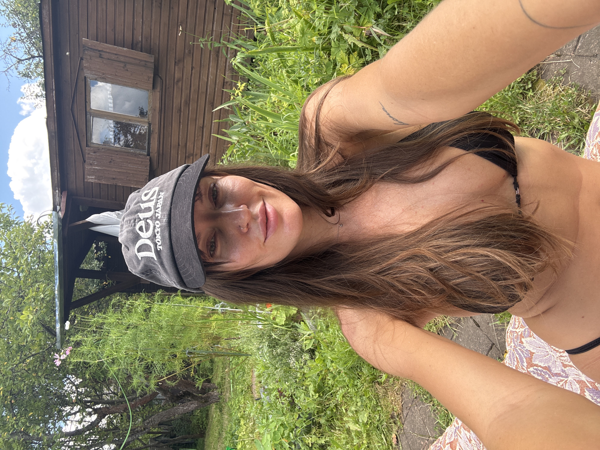 The image shows a woman taking a selfie in a garden. She is wearing a black bikini top and a grey baseball cap with "DEUS TOKYO JAPAN" printed on it. Her long brown hair is flowing down her shoulders. She has a tattoo on her arm. The background features a wooden shed with a small window, lush green foliage, and trees. Sunlight suggests it is daytime, and the greenery indicates a warm, possibly summer, season. The setting appears to be a rural or semi-rural area, consistent with a "SNT" (Sadovodcheskoye Nekomercheskoye Tovarishchestvo, or gardening non-profit partnership) in Russia. The woman's expression is relaxed and natural, as if she is enjoying a sunny day outdoors.