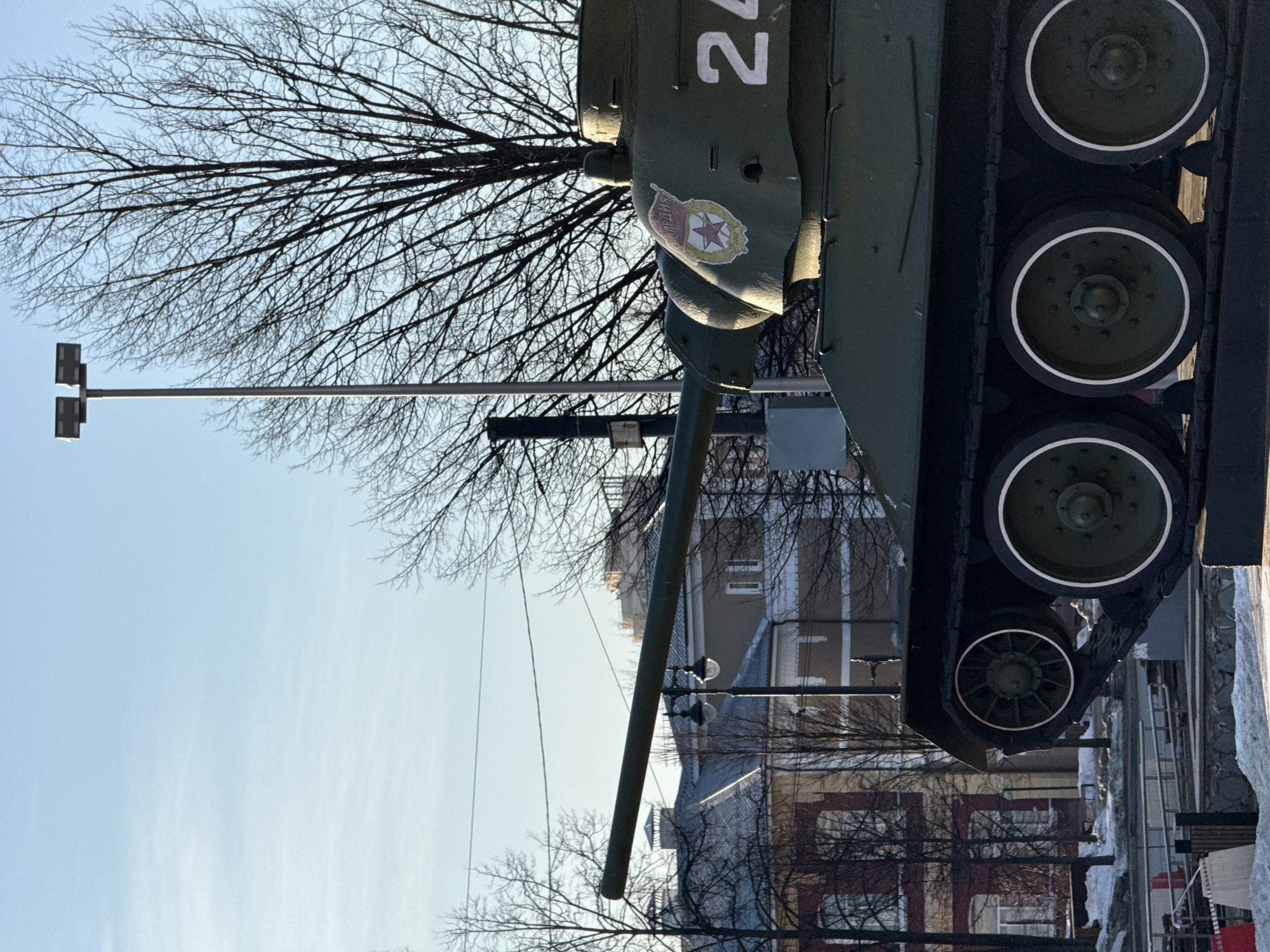 The image displays a green Soviet-era T-34 tank monument, observed from its right side, with the main gun barrel aimed leftward. The tank's turret features a circular red and yellow emblem containing a red star and the Cyrillic word "ГВАРДИЯ" (Guard), alongside the white painted number "24". The tank is mounted on a dark, elevated base. In the background, under a light blue sky with minimal cloud cover, numerous leafless deciduous trees are visible. Multi-story buildings, some with reddish-brown and others with beige facades, are present in the mid-ground. A tall, grey metal street light pole with two rectangular luminaires stands behind the tank, and an older black lamppost with two globular lights is positioned behind the tank's barrel. Patches of snow are visible on the ground in the foreground, alongside a concrete barrier with red and white markings and metal railings. No human figures are present. This monument is located at Komsomolsky Prospekt in Perm, Russia.