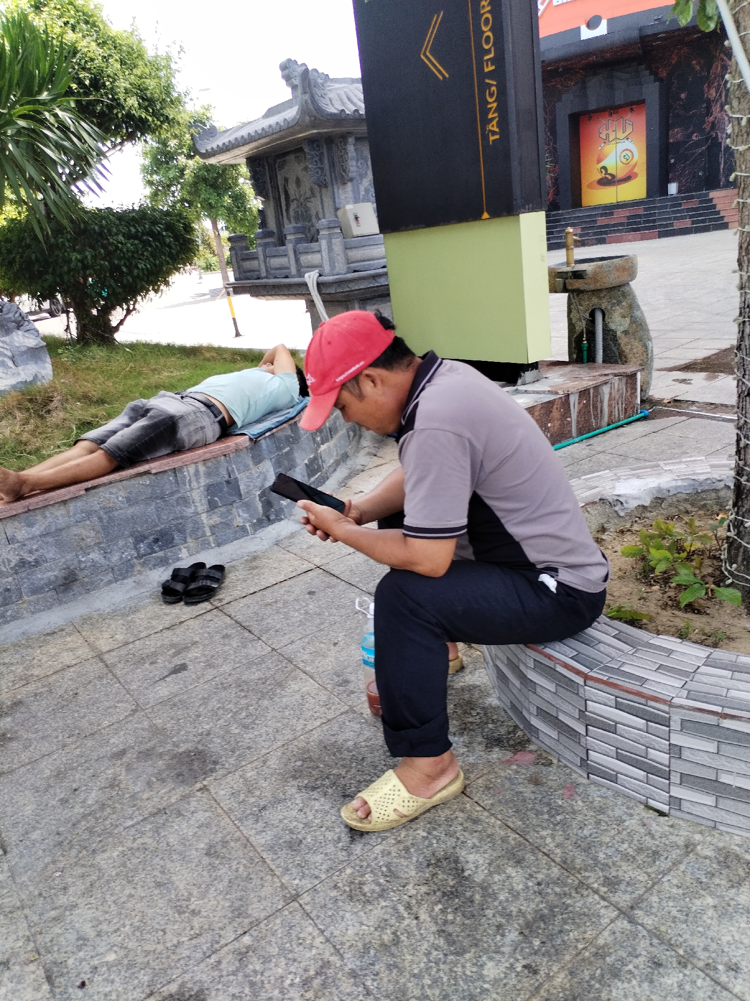 In Nha Trang Ward, Vietnam, a man wearing a red cap and a grey t-shirt sits on a tiled bench, looking down at his phone. His dark pants are rolled up, and he wears light yellow sandals. Beside him, a person wearing a light blue t-shirt and grey shorts lies on their side on a stone ledge, with their head on a blue mat and their hands behind their head. A pair of black sandals rests on the ground near the ledge. In the background, lush green trees and a stone structure with intricate carvings are visible, along with a sign that reads "TANG / FLOOR". The scene is set on a paved area with a patterned tiled bench.