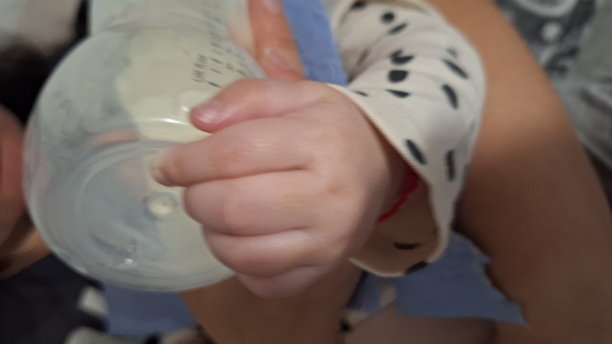 A baby's hand grips a clear plastic baby bottle filled with a milky liquid. The baby's arm, clothed in a light-colored long-sleeved garment with dark spots, is visible. A red string is tied around the baby's wrist. A second adult arm is visible in the background, and there is an out-of-focus view of a person's face on the left side of the picture. The background appears to be a blurry, indeterminate indoor setting. There is no information in the photo to suggest the location of Blagoevgrad, Bulgaria.