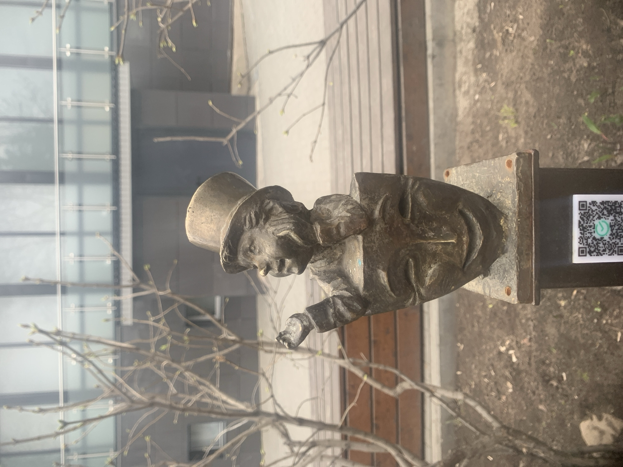 The image depicts a bronze bust of a person wearing a top hat. The bust is mounted on a dark post with a square base. A QR code is affixed to the side of the post. Bare branches with emerging buds are visible in the foreground, partially obscuring the bust. In the background, there's a modern building with reflective glass windows and a wooden-clad wall.
Based on the details, this is likely the bronze bust of Izhevsk's 'Sokolyonok' located near the National Library in Izhevsk, Russia.