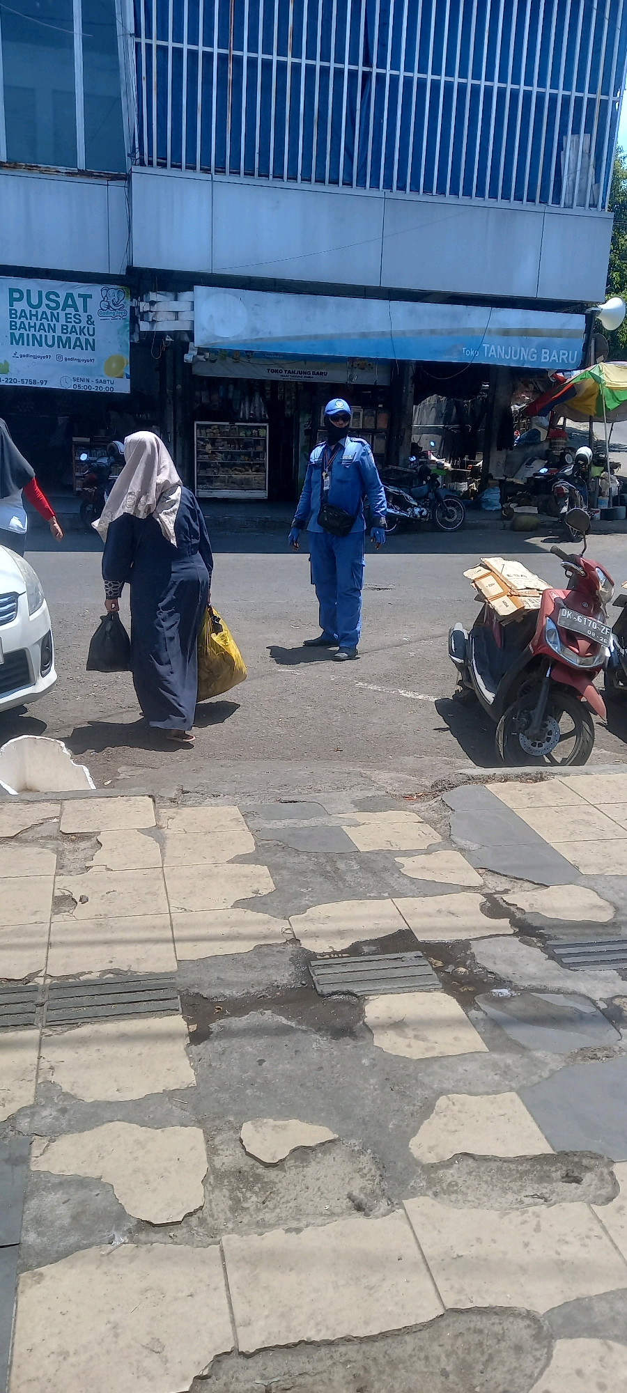 In Jember, Indonesia, a street scene unfolds with a focus on pedestrian and vehicular activity. A person dressed in a bright blue uniform, possibly a security guard or maintenance worker, stands near a crosswalk, wearing a cap and face mask, and a dark bag is slung across their chest. To the left, a woman in a dark abaya and a light beige hijab walks away from the camera, carrying two bags, one black and one large yellow, in her hands. A white car is partially visible on the far left. In the background, shops line the street, with signs indicating "PUSAT BAHAN ES & BAHAN BAKU MINUMAN" (Center for Ice and Beverage Raw Materials) and "TOKO TANJUNG BARU" (Tanjung Baru Store). Several motorcycles are parked or visible on the street, including a red motorcycle in the foreground with a license plate visible. The pavement in the foreground appears worn and damaged, with patches of exposed concrete and scattered debris. The environment suggests a typical urban street in Indonesia, with a mix of commercial establishments and everyday life.