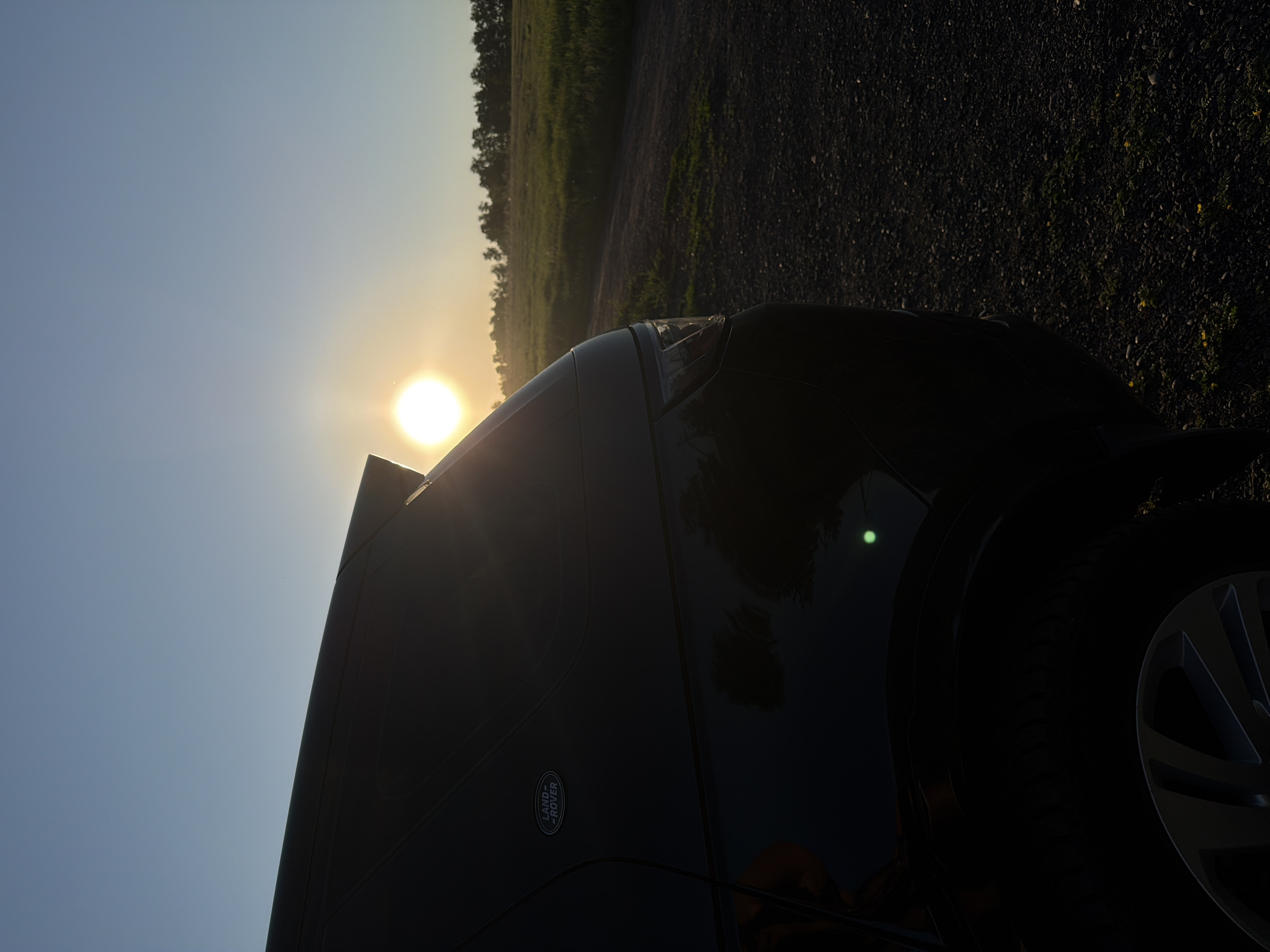 This image, originally presented in a portrait orientation but depicting a landscape scene, features a dark-colored Land Rover vehicle, likely an SUV, parked on a gravel road. The vehicle is visible from its left side and front, with the "LAND ROVER" logo clearly discernible on its side panel.
The sun is low on the left horizon, casting a powerful, warm light across the scene, indicating either sunrise or sunset. This backlighting creates significant lens flare and bright highlights on the vehicle's roofline and windshield, while much of the vehicle and the immediate foreground remain in shadow. To the right of the vehicle, a green field stretches towards a line of trees in the distance. The sky transitions from a brilliant, sunlit expanse near the horizon to a clear blue overhead.
No individuals or specific activities are depicted, contributing to a serene outdoor ambiance. The setting appears to be a rural or semi-rural area, consistent with the provided location context of Krasnoyarsk, Russia.