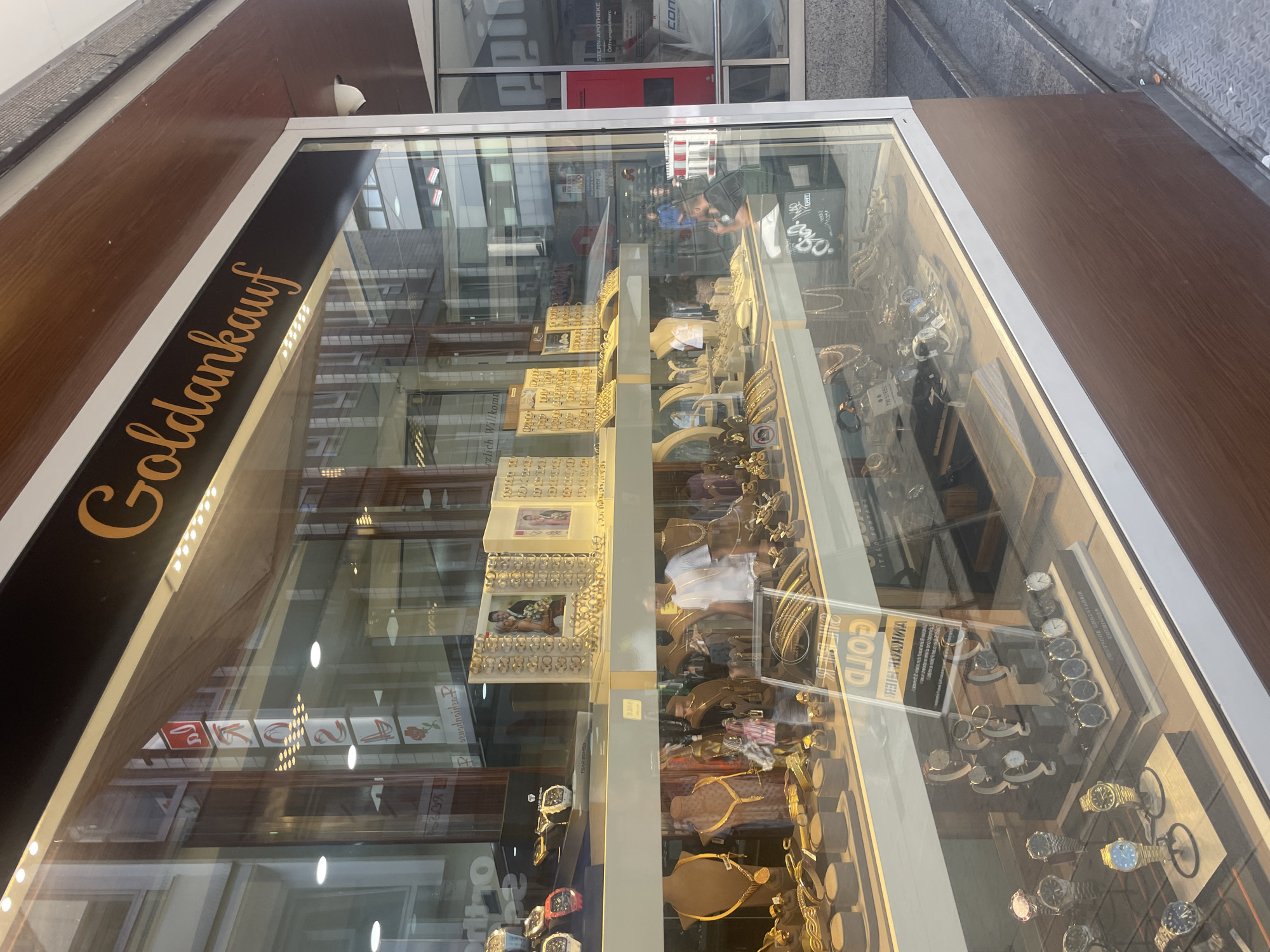 The image shows the display window of a jewelry store called "Goldankauf" in Wuppertal, Germany. The store's name is written in orange cursive lettering on a dark background above the window. The display window is filled with various jewelry items, including necklaces, bracelets, rings, and watches, all meticulously arranged on shelves and mannequins. In the background, the reflections in the glass reveal buildings and street scenes, indicating an urban setting. A sign inside the store, partially visible, reads "Goldankauf" and also includes "Ankauf Hier," suggesting they buy gold. There are no people clearly visible inside or outside the store. The lighting appears to be a mix of interior store lights and natural daylight.