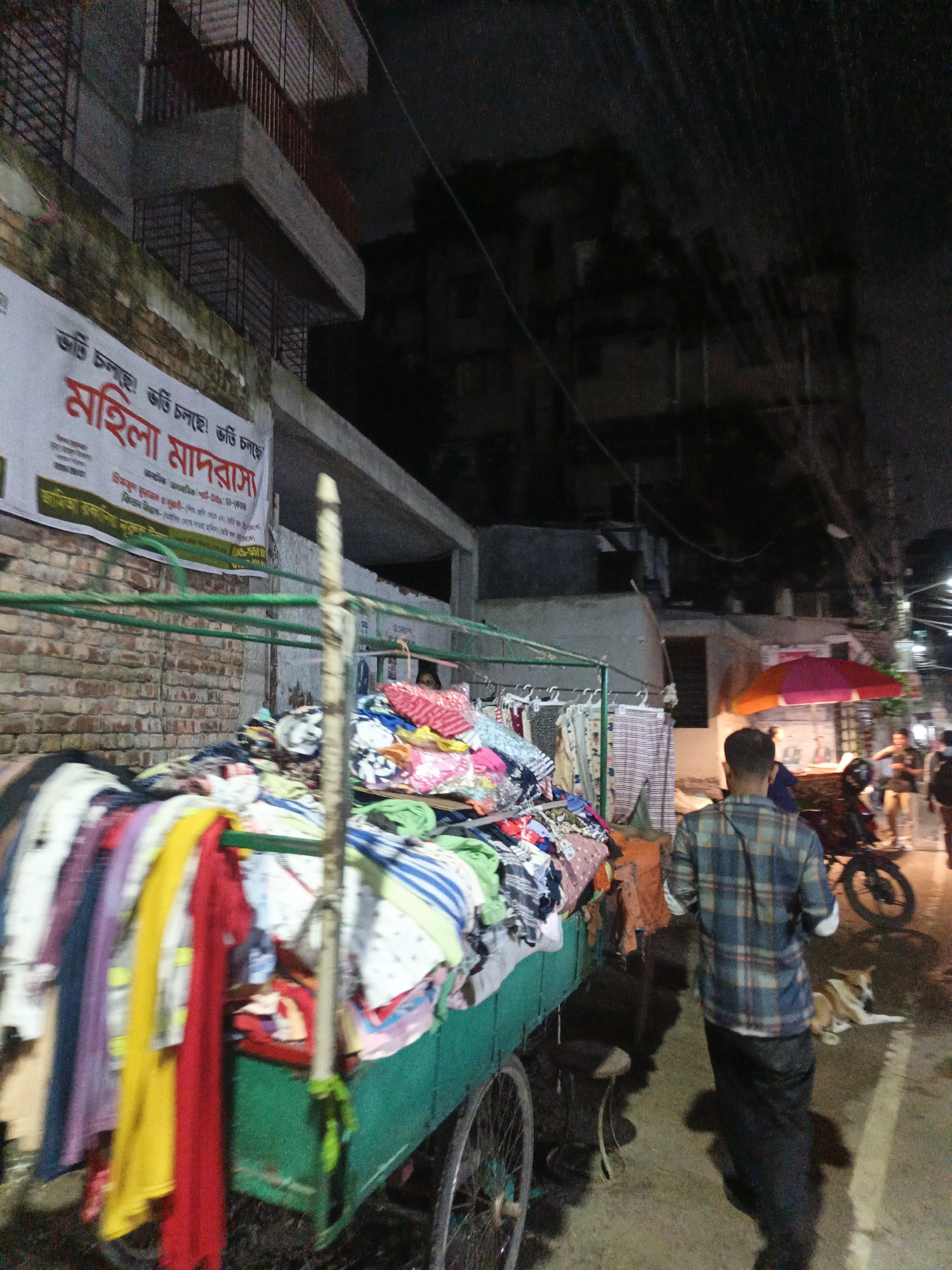 The image depicts a street scene at night in Dhaka, Bangladesh, near a building with a brick facade. A large cart, likely a street vendor's stall, is filled with an assortment of colorful clothing. The cart is green and has metal racks holding more garments. A man wearing a plaid shirt and dark pants is walking away from the camera on the wet pavement. To the right, a motorcycle is parked, and a dog is lying on the ground. In the background, there is a pink and orange umbrella over a stall and other indistinct figures and structures, suggesting a busy urban environment. A banner with red text is visible on the brick wall. The overall impression is that of a lively, perhaps slightly chaotic, street market at dusk or night.