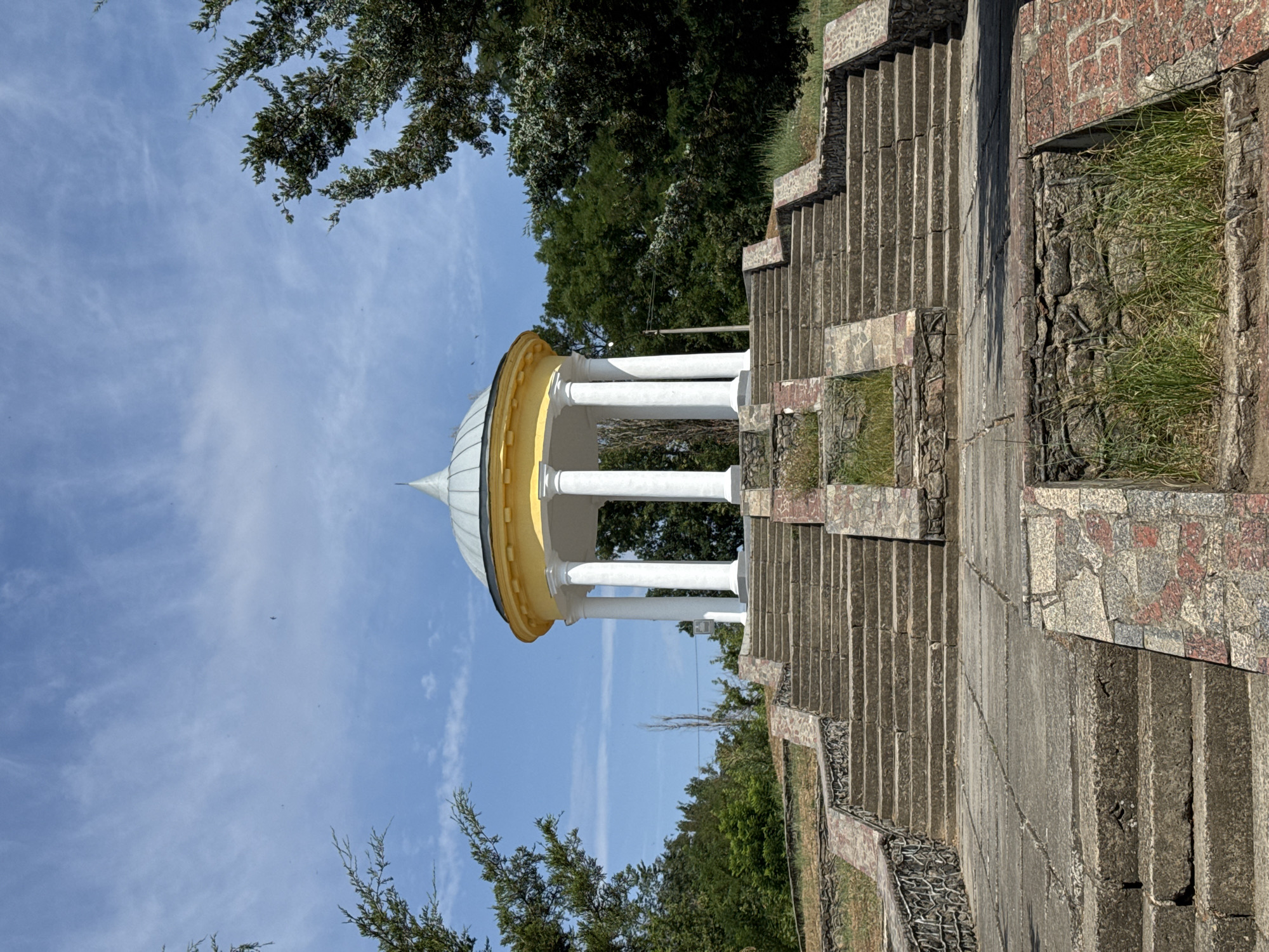 The image depicts a monument with a circular, yellow-roofed pavilion supported by white columns, situated atop a stone staircase. The pavilion has a conical, silver-colored roof. The staircase, constructed from concrete and adorned with stone accents, ascends towards the pavilion. The surrounding environment is characterized by lush green trees and a clear blue sky with wispy white clouds. The scene is located in the city of Voznesensk, Ukraine, at the historical monument dedicated to the victims of political repressions. There are no people visible in the image.
