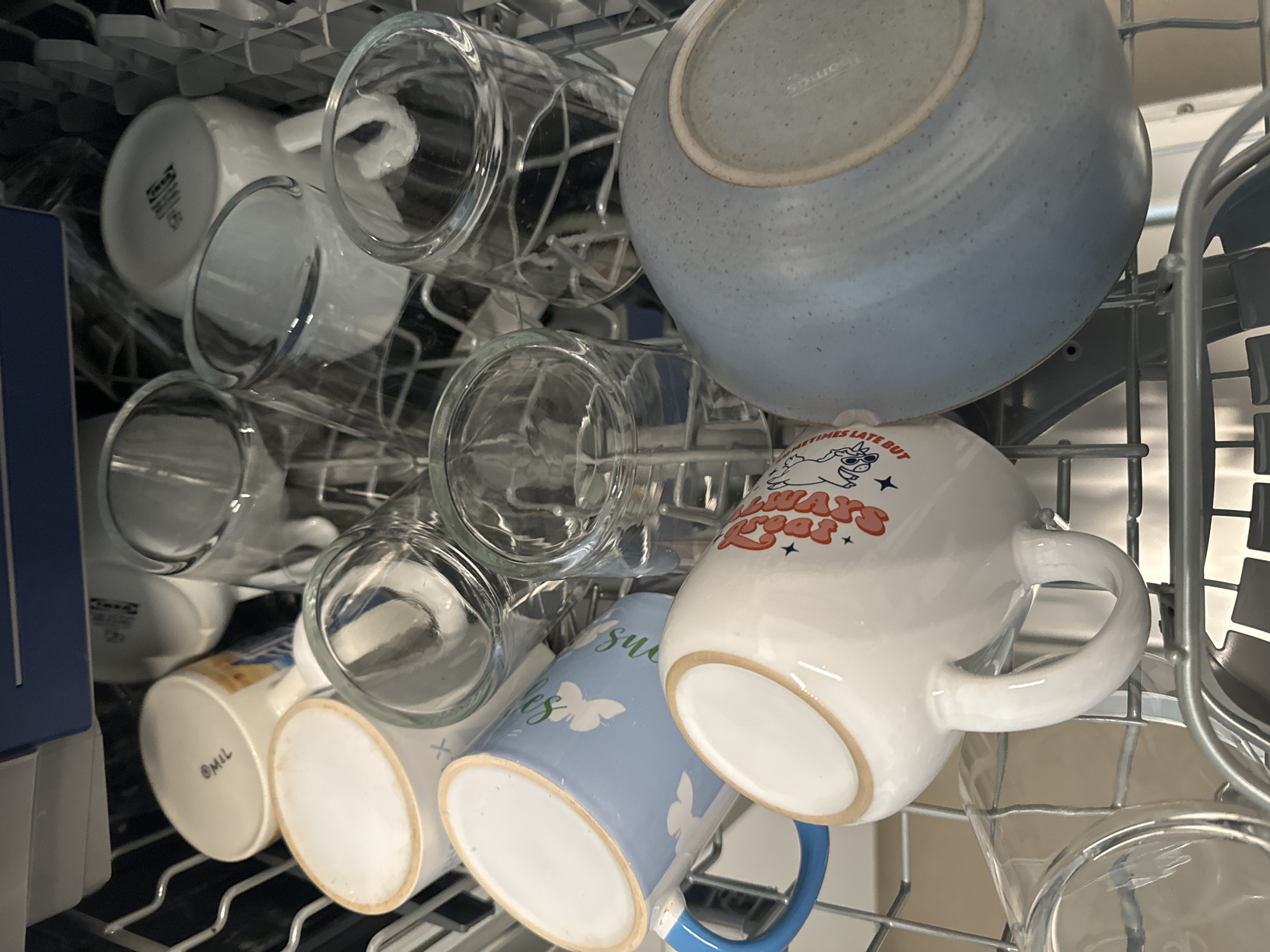 The image shows a close-up view of dishes in a dishwasher rack. The primary subjects are various mugs and glasses, predominantly made of glass and ceramic. A light blue speckled bowl is also visible, resting on top of the rack. The scene is the interior of a dishwasher, indicated by the metal racks and the overall organized placement of the dishware.
The activity taking place is the loading or unloading of a dishwasher, or the dishes being washed. The lighting suggests an indoor setting, and the overall tone is neutral, focusing on the objects themselves.
Notable details include the text visible on some of the mugs. One white mug has red lettering that reads "ALWAYS LATE BUT". Another blue mug has a white butterfly design and some green text that is partially obscured but appears to say "sweet". A white mug on the left has text printed on its base, part of which reads "@Mil". The dishes are arranged in a typical dishwasher configuration, with glasses upright in their slots and mugs placed between them.
There are no visible cues about the time of day or weather. The location context of Murcia, Spain, does not provide specific visual information within the frame itself. The image is focused on the domestic setting of a kitchen appliance.