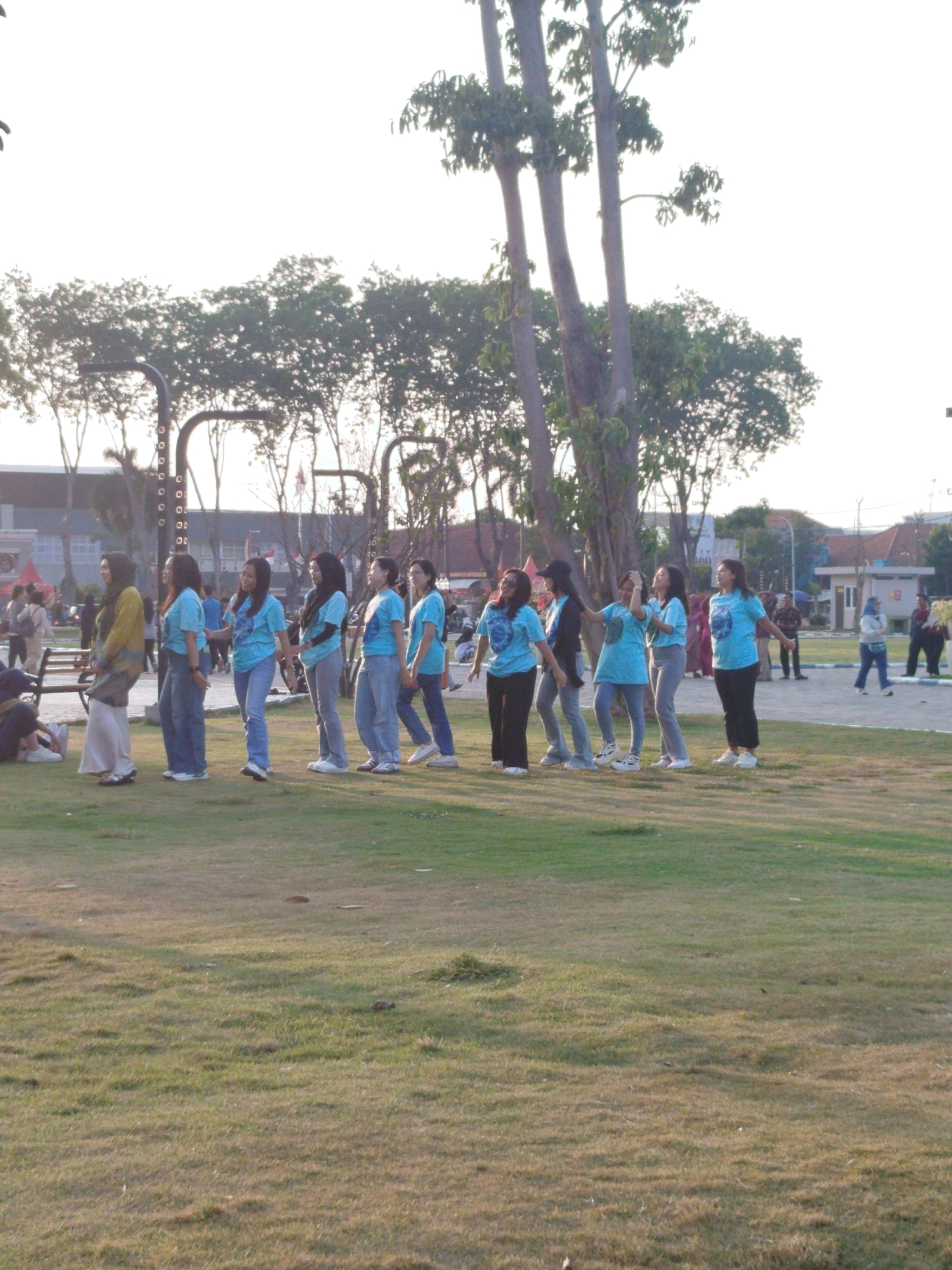 In Sidoarjo, Indonesia, a group of women in blue t-shirts and jeans stands in a grassy park. The women are lined up in a row, facing left, and appear to be engaged in a coordinated activity. They are all wearing similar outfits, suggesting they are part of a group or team. The park is landscaped with trees and a paved path, and in the background, buildings and more trees are visible. The sun is low in the sky, casting a warm glow on the scene.