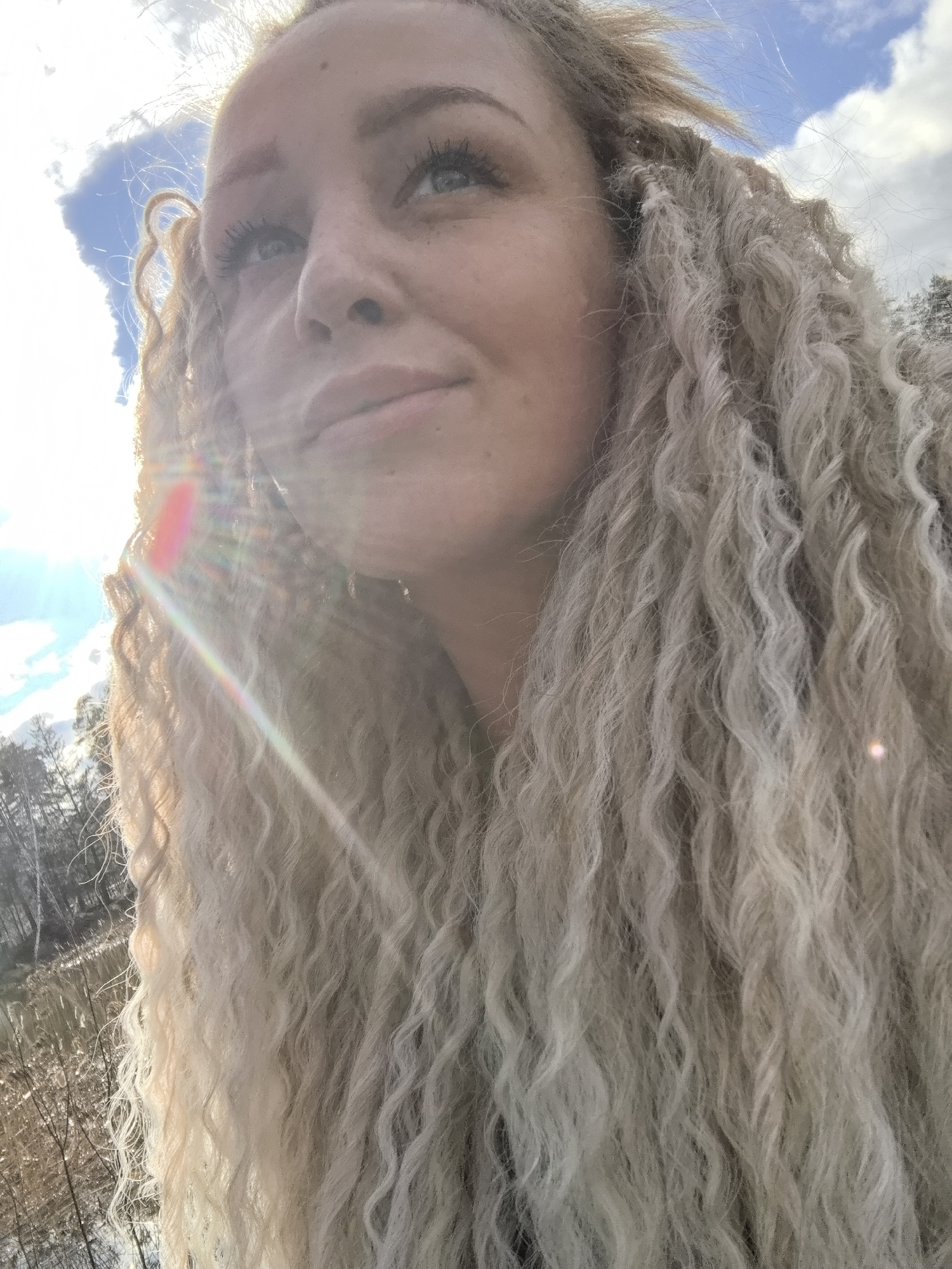 A close-up image features a fair-skinned individual with blond, curly hair. The person is looking upward, with a slight smile. The sky is visible in the background with some clouds and what appears to be sunlight. Trees and water are visible in the background. It is not possible to confirm whether the picture was taken in the city of Noviy, Russia.