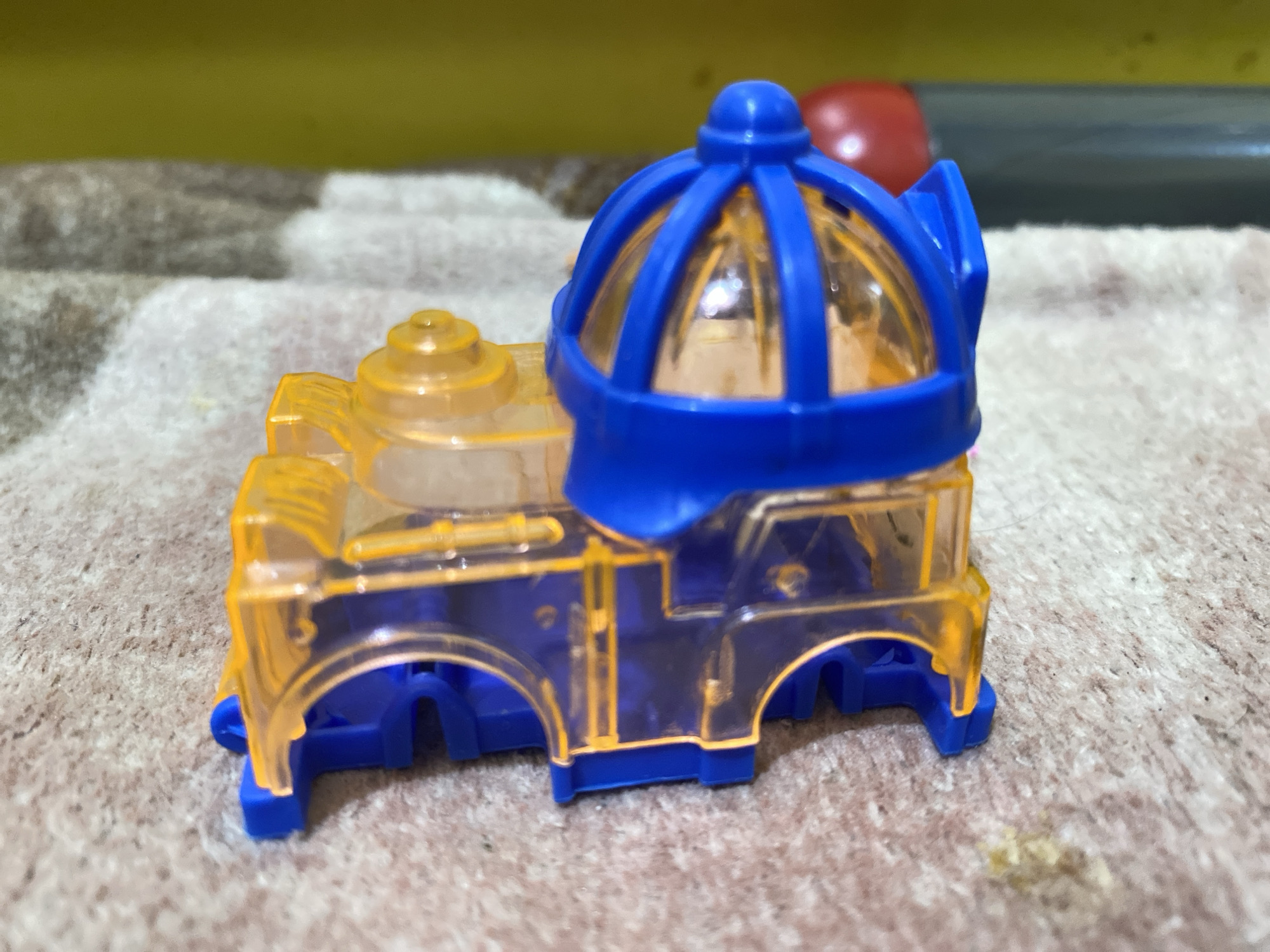 A translucent orange toy train, with a blue dome-shaped top and blue wheels, sits on a textured surface. The background is blurred, showing yellow and brown colors. The scene is devoid of any human presence. The toys are not in Lambaro Kueh, Indonesia.