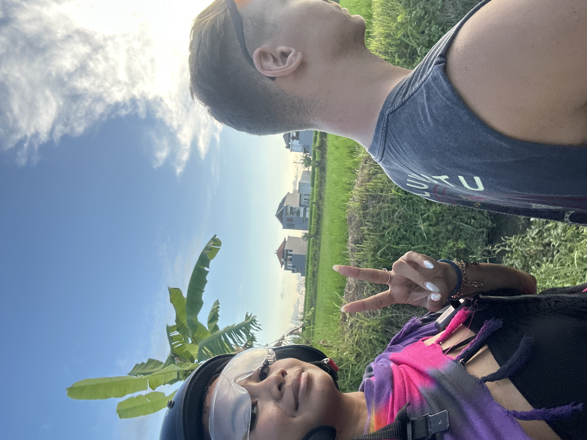 A selfie taken outdoors in Sukawati, Indonesia, showing two individuals. One person is wearing a black helmet and transparent goggles, with a bright tie-dye scarf/shawl and giving a "peace" sign with their hand. The other person is wearing a grey tank top and sunglasses on their head, looking towards the sky. The background includes a partly cloudy blue sky, rice paddies, and houses in the distance. A banana tree is on the left.