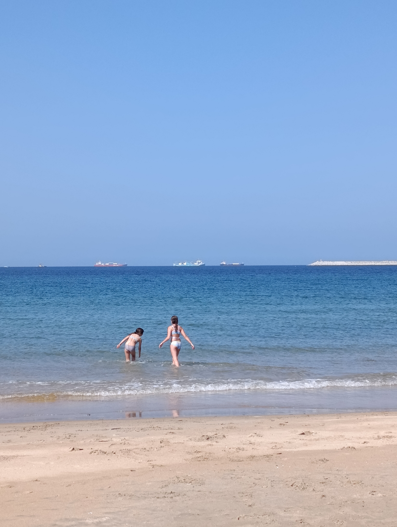 The image depicts a sunny day at a beach, likely in Sines, Portugal. Two young individuals, one appearing to be a child and the other a teenager, are standing in the shallow water at the edge of the sea. The child, facing away from the camera, is bent slightly, possibly playing or looking at something in the water. The teenager, also facing away, is standing upright with arms extended slightly to the sides. Both are wearing swimsuits.
The ocean is calm with gentle waves lapping the sandy shore. In the background, several cargo ships are visible on the horizon, indicative of a busy shipping lane. The sky is clear and a vibrant blue. The beach itself is composed of light-colored sand, with some debris scattered along the waterline. The scene is peaceful and suggests a recreational outing at the coast.