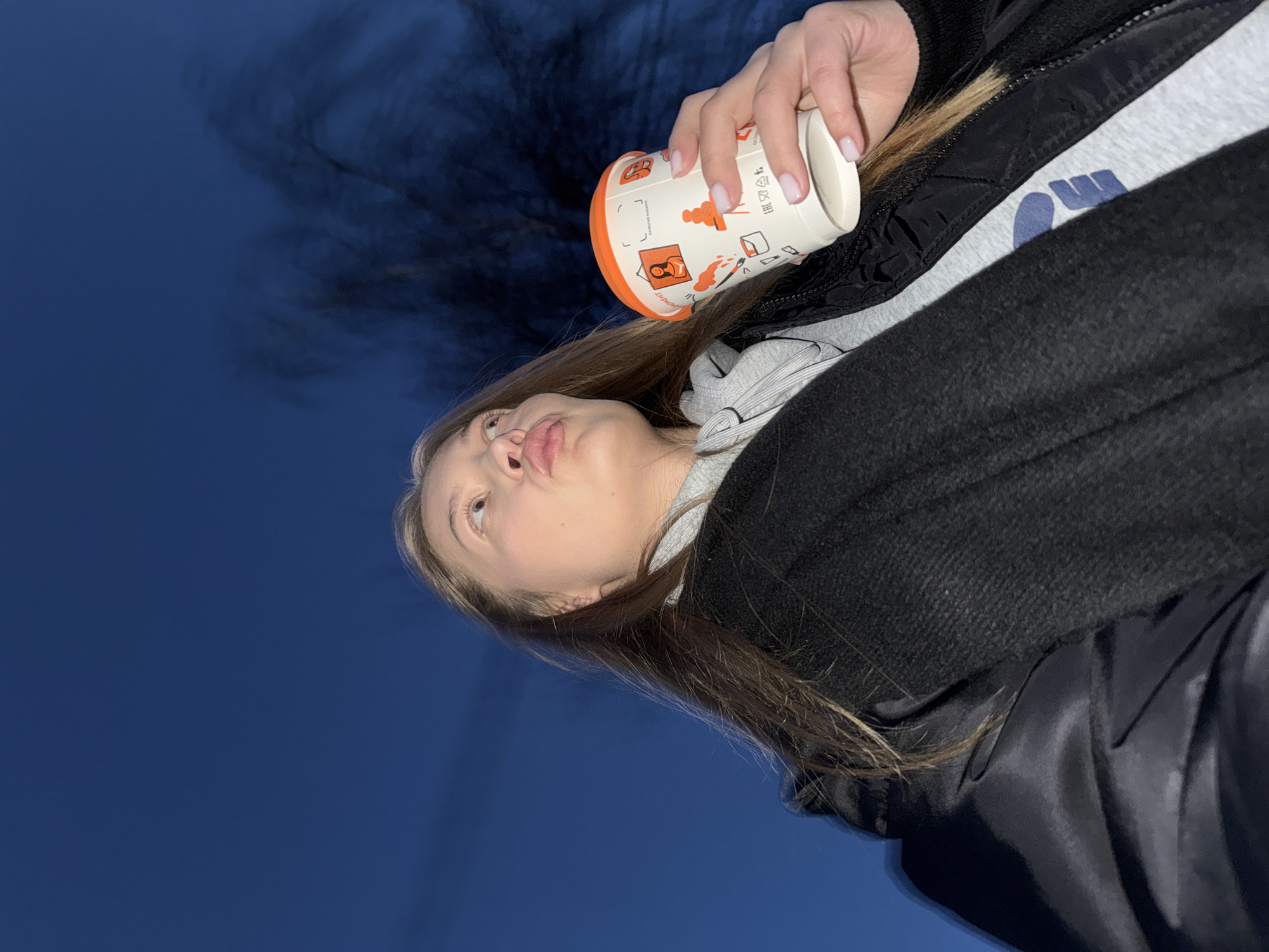 A young, light-skinned woman is shown in a low-angle, rotated perspective, holding a white paper cup with orange decorative images near her face. She is wearing a grey hoodie with a blue logo, underneath a black jacket. The background is a dark blue, possibly the night sky or a dark building. It is unknown if the location is in the city of Chaikovsky, Russia, as there are no identifying landmarks.