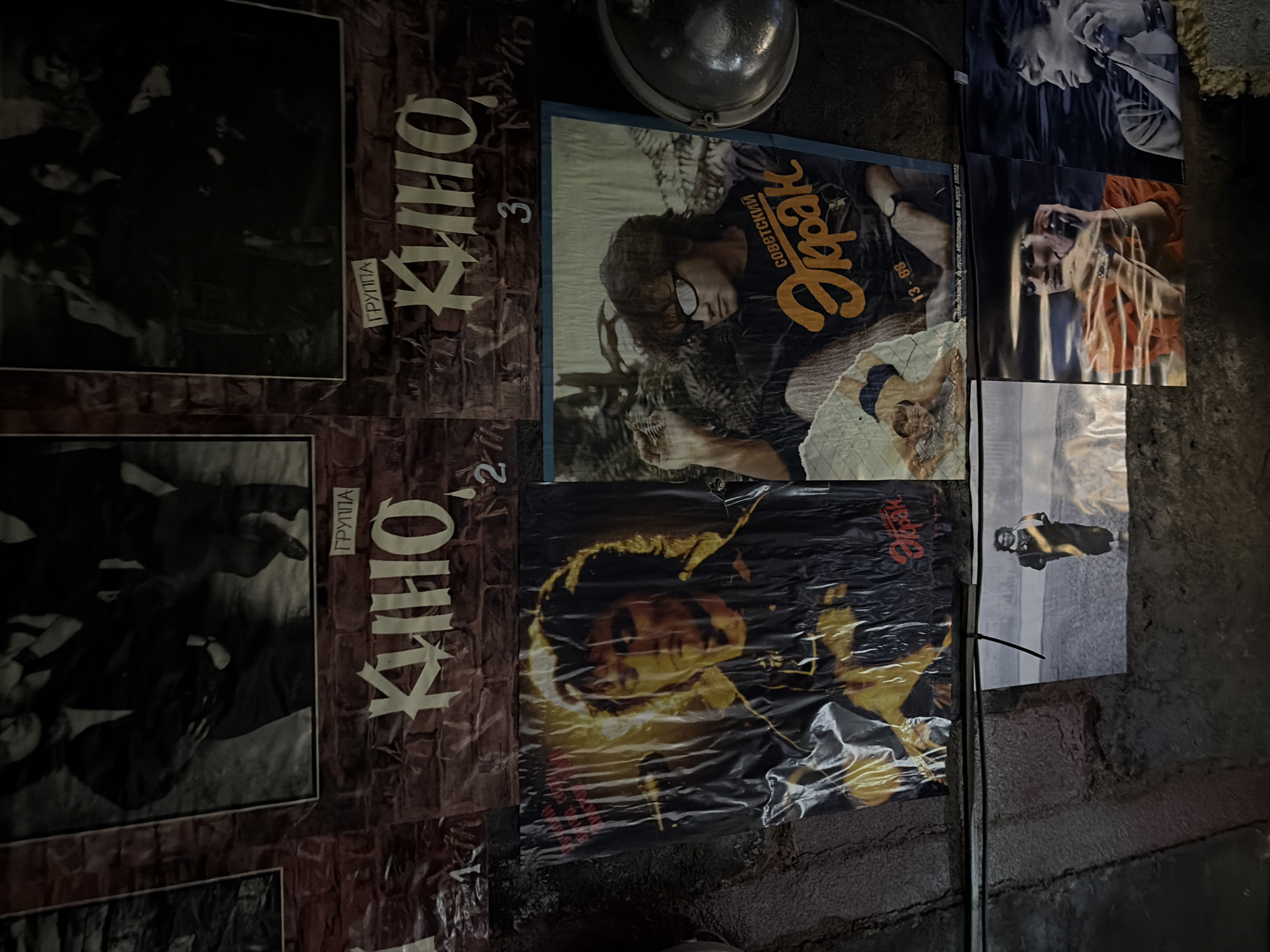 The image depicts a wall adorned with posters and photographs, likely in Saint Petersburg, Russia, given the presence of posters for the band "Кино" (Kino). The wall itself appears to be made of brick or a similar textured material, with a rough, aged appearance.
Dominating the upper portion of the image are two large posters featuring black and white photographs of what appear to be band members. Text above these posters reads "ГРУППА" (Group) followed by "КИНО" in large, stylized Cyrillic lettering. Numbers "1", "2", and "3" are visible below the "КИНО" lettering, suggesting a possible ranking or a series.
Below these, two prominent posters showcase individuals. The poster on the left features a close-up portrait of a man with wavy hair, illuminated by yellow light, and wearing dark clothing. The poster on the right displays a man wearing sunglasses and a t-shirt with the text "СОВЕТСКИЙ Экран" (Soviet Screen) and the date "13-08".
Further down, a series of smaller photographs are arranged. One photograph shows a person standing in a desolate, possibly outdoor landscape. Another poster features a person in an orange top, appearing to be on the phone. To the right, a black and white portrait of a man's face is visible.
A metallic circular object, possibly a lamp or part of a fixture, is visible on the far right of the image. The overall lighting is dim, with some posters reflecting light. The environment appears to be an indoor space, possibly a public area or a collection space dedicated to the band Kino.