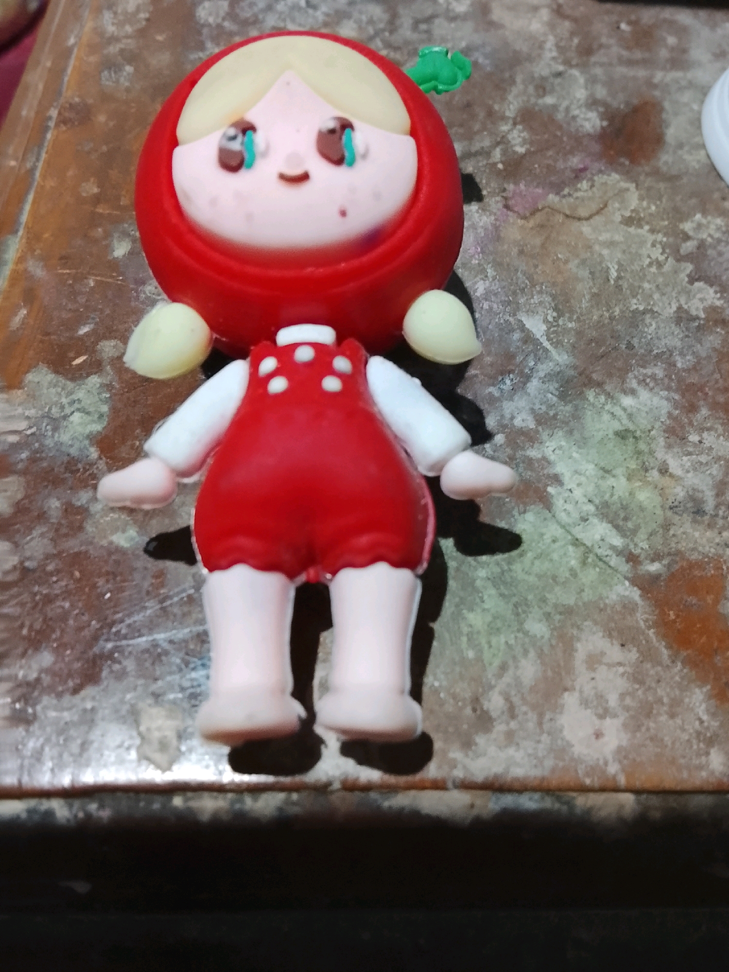 A handmade doll, approximately 6 inches tall, is centered on a rough, textured wooden surface. The doll has a round red head with blonde hair peeking out. Its face is pink with two large brown eyes outlined in black and blue irises, and a small curved mouth. A tiny green sprout is attached to the top of its head. The doll's body is wearing a red dress with white polka dots and a white collar. It has thin white arms and light pink hands, and light pink legs with round feet. The wooden surface appears worn and possibly aged, with visible grain and some lighter-colored discoloration. The lighting casts a distinct shadow of the doll onto the surface. The doll is positioned upright, as if standing. There is no visible indication of the doll's location within the city of Ujungrusi, Indonesia.