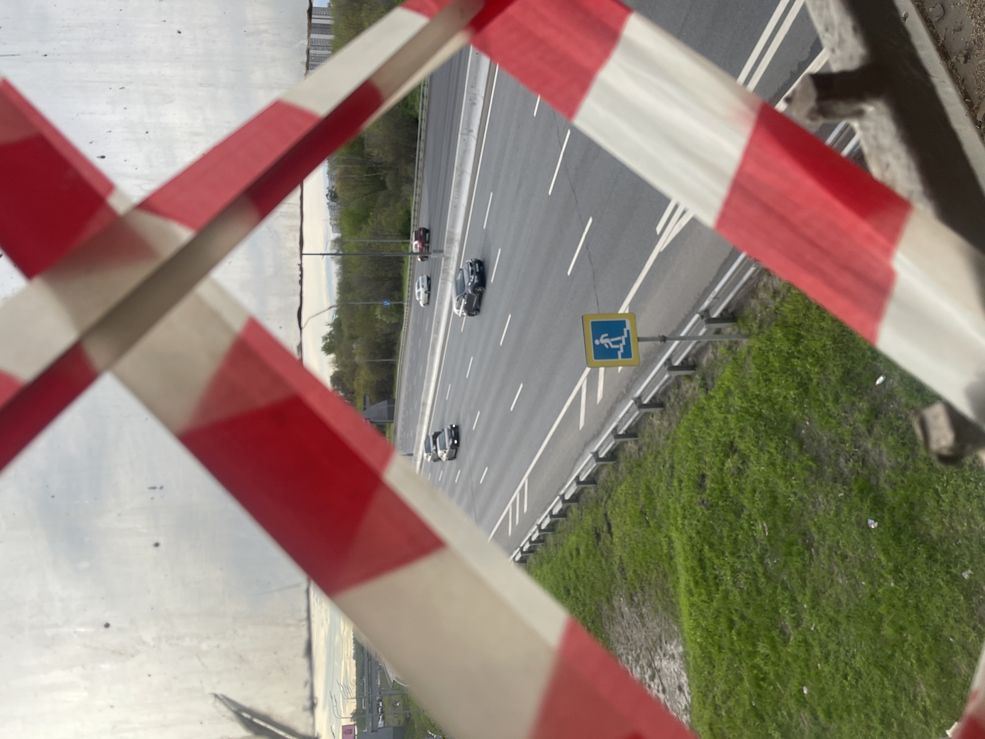 The image depicts a view of a highway in Вёшки, Russia, as seen through a barrier marked with red and white stripes. Several vehicles are visible on the highway. A pedestrian crossing sign is positioned alongside the road. Green grass covers the ground next to the highway.