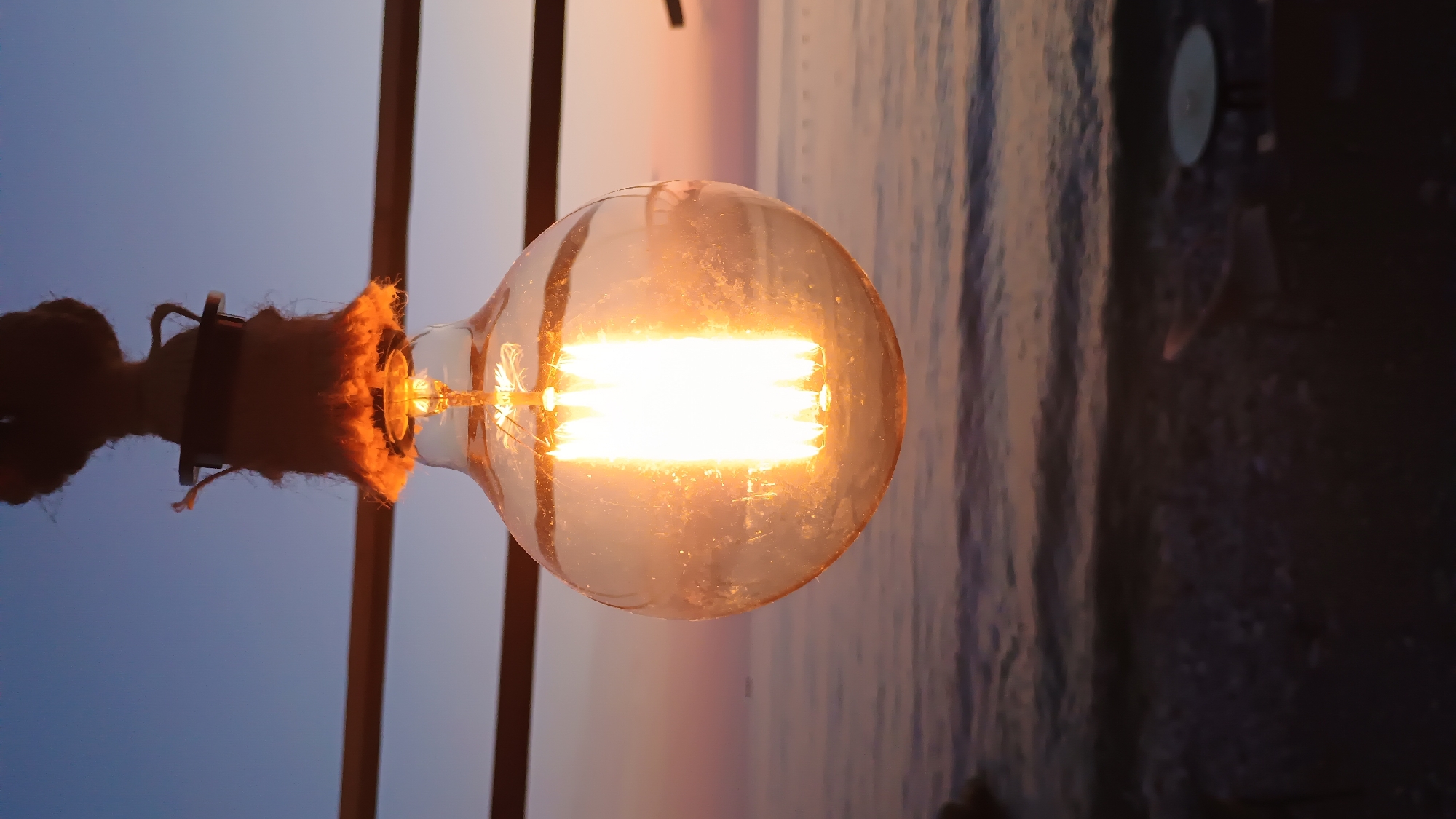 A close-up, horizontal, and outdoor shot depicts an illuminated Edison-style light bulb with a clear glass bulb and visible filaments, suspended by a rope-wrapped fixture. The background reveals a scene suggestive of a coastal environment with the sky transitioning from blue to hues of pink and orange, typical of a sunset or sunrise. The sea or ocean water appears rippled and reflects the colors of the sky. The light fixture is attached to a dark, vertical pole or structure. The precise location is unidentifiable.