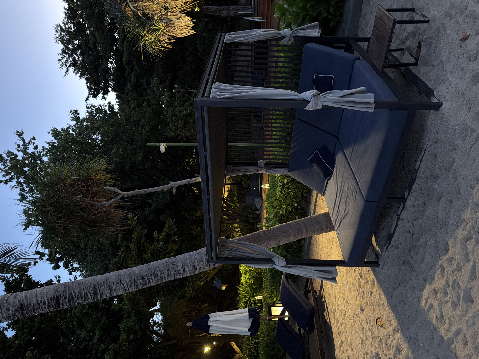 The image depicts a beachside scene in Ko Samui District, Thailand, during dusk. The foreground features a dark blue, two-person cabana bed with grey curtains tied back, set on a sandy beach. Adjacent to the cabana is a small wooden side table. In the background, several dark blue lounge chairs are visible, along with a closed beach umbrella. The environment is tropical, characterized by lush green foliage and palm trees, with some ambient lighting casting a warm glow. The sky is a deep blue, indicating late evening. No people are present in the scene.