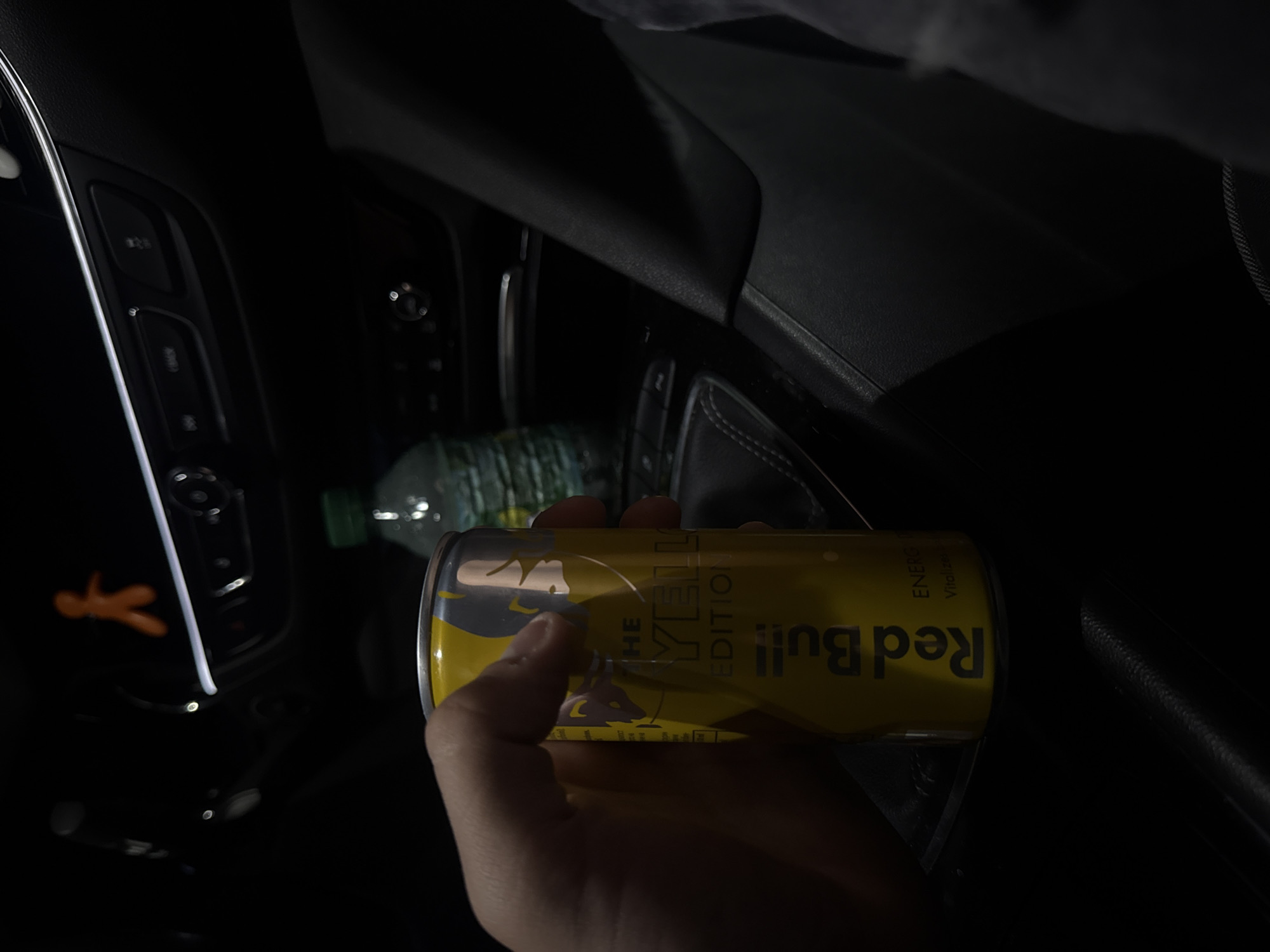 An interior shot depicts a human right hand holding a yellow Red Bull "The Yellow Edition" energy drink can within a vehicle, specified as being in Varna, Bulgaria. The can is primarily yellow with silver accents and text, held firmly by the thumb and fingers. In the background, the dark interior of a car is visible, featuring a dashboard with illuminated controls and a prominent horizontal chrome trim strip. An out-of-focus orange, human-shaped air freshener or decoration is affixed to the upper dashboard. To the right of the Red Bull can, a green-capped plastic water bottle is partially visible and out of focus. The lower right portion of the image shows dark vehicle upholstery and components, possibly part of the center console or passenger seat area. Lighting is dim, indicating low-light conditions, possibly at night. No other individuals are visible.
