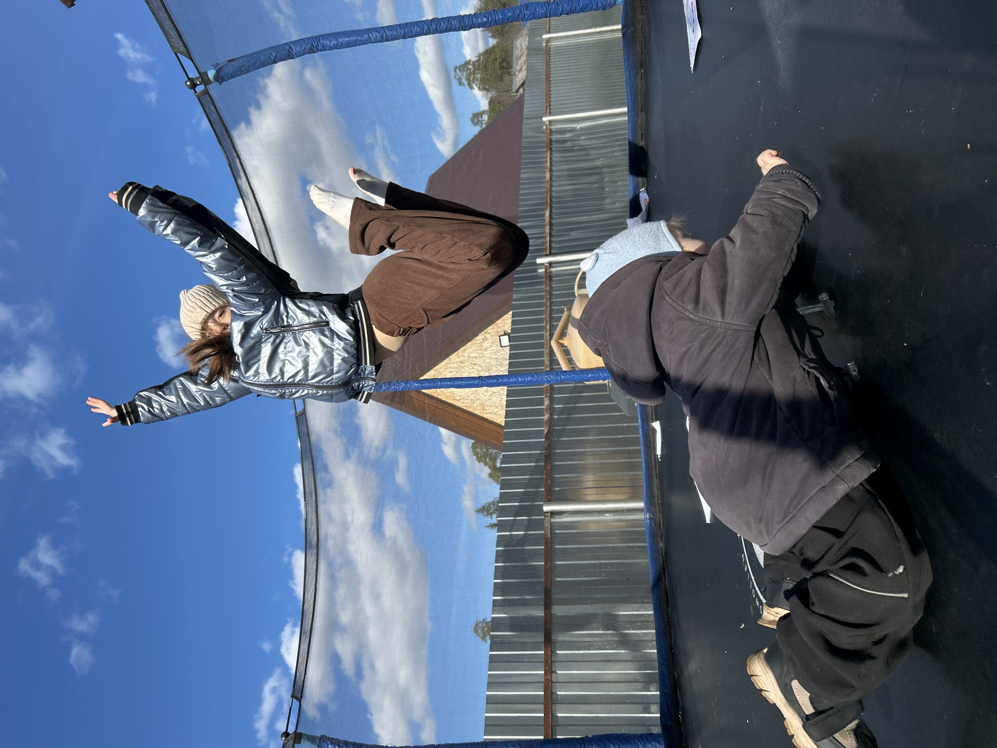 The image depicts two individuals on a trampoline, likely taken in Chaikovsky, Russia, given the request. One person is airborne, suspended against a backdrop of a bright blue sky with scattered clouds. This individual is wearing a silver-toned jacket, brown pants, and a light-colored beanie, with arms outstretched. The second individual is on the trampoline surface, wearing a dark-colored jacket, dark pants, light-colored shoes, and a light-colored hat. The trampoline is enclosed by a safety net. In the background, a roof is visible, along with a corrugated metal fence. The environment appears to be outdoors, under daylight conditions.