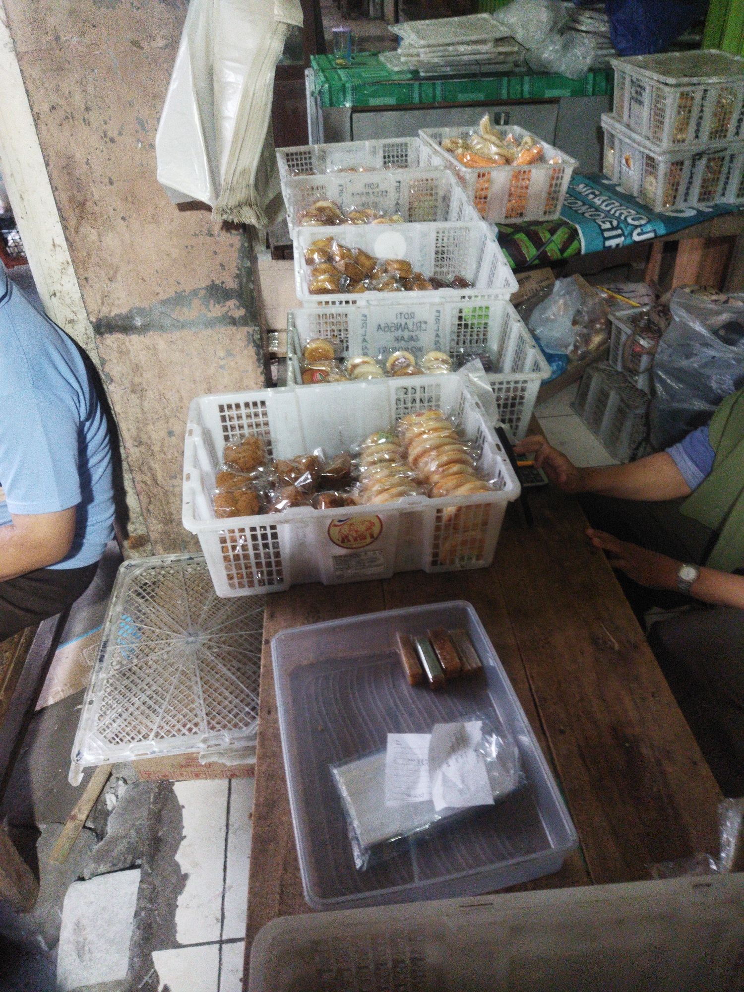 This image depicts a market stall, likely in Wonogiri, Indonesia, with a variety of baked goods displayed in white plastic crates. In the foreground, a person wearing a blue shirt and dark pants is partially visible on the left, seated. To the right, another person, wearing a green shirt and dark pants, is seen from the chest down, with their hands interacting with a calculator. Several white plastic crates are stacked and arranged, filled with pastries and bread, some of which are individually wrapped in plastic. A plastic tray containing wrapped food items and paper slips sits on a wooden table in the lower right portion of the image. The background shows an unpainted, rough-textured wall and other market elements like stacked crates and bags. The ground appears to be tiled and cracked in places.