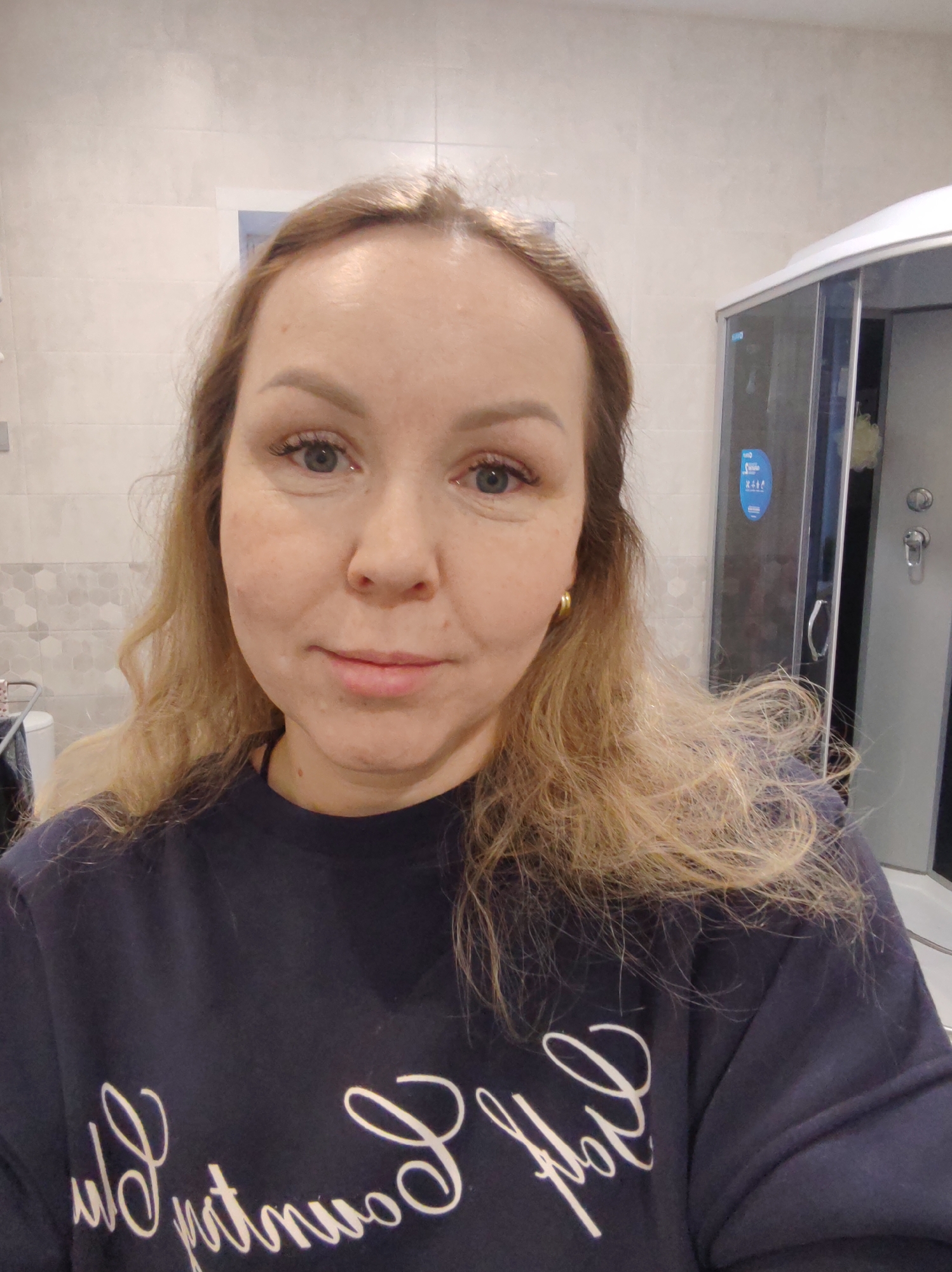 The image displays a woman taking a mirror selfie in a bathroom located in the city of Chaikovsky, Russia. The subject, a woman with light-colored eyes and shoulder-length, wavy, light brown to blonde hair, gazes directly at the camera. She is wearing a dark-colored long-sleeved top with white script lettering, which appears reversed due to the mirror, partially spelling "y Country" or a similar phrase. A single gold hoop earring is visible on her right ear. The bathroom walls are tiled with light grey rectangular tiles on the upper section and a horizontal band of grey hexagonal patterned tiles at mid-height. To the right, a modern shower cabin with clear glass panels, a silver frame, and a visible chrome mixing valve is present. Inside the shower, a yellow-green loofah or sponge hangs. To the left, a white toilet and a metal drying rack are partially visible behind the woman's left shoulder.