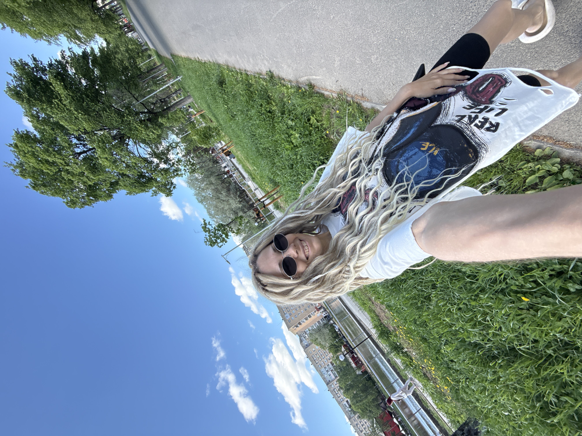 A tilted outdoor selfie depicts a person with blonde dreadlocks and sunglasses, smiling at the camera. They are wearing a white t-shirt with a graphic print. Part of their body is visible, clad in black leggings and white sandals. They are holding a white graphic tote bag. The background includes a blue sky with white clouds, green trees and grass, and buildings in the distance. A paved path is visible in the background. The image appears to be taken near a park or green space within a city, the location of which is unknown.