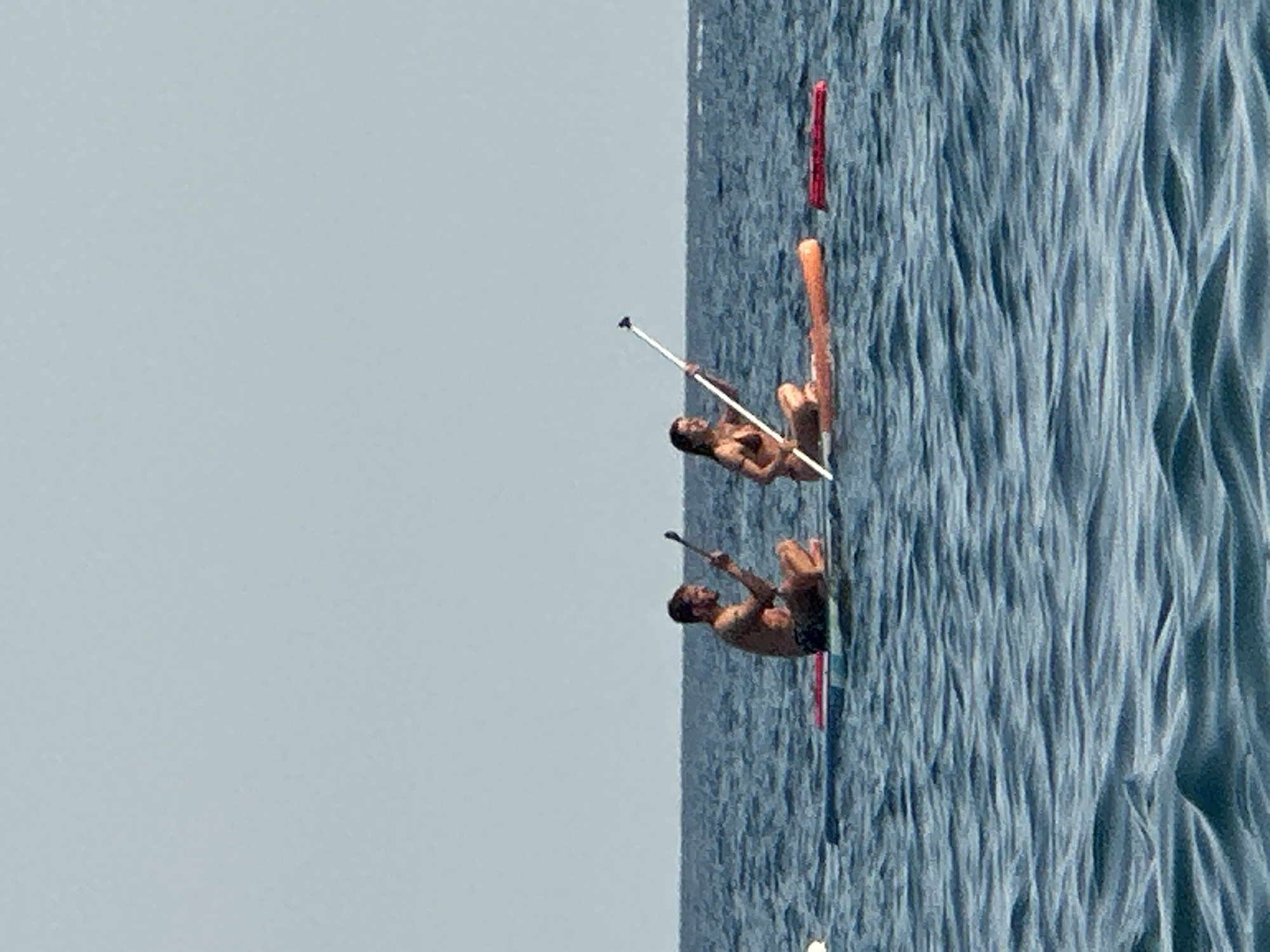Two individuals are paddleboarding on a body of water. The person on the left, appearing to be male, is seated and holding a paddle. The person on the right, appearing to be female, is also seated and holding a paddle. Both are wearing swimwear. They are on separate paddleboards. The water is choppy with visible waves. The sky is clear and pale blue. This scene is occurring in the waters off the coast of Boreti, Montenegro.