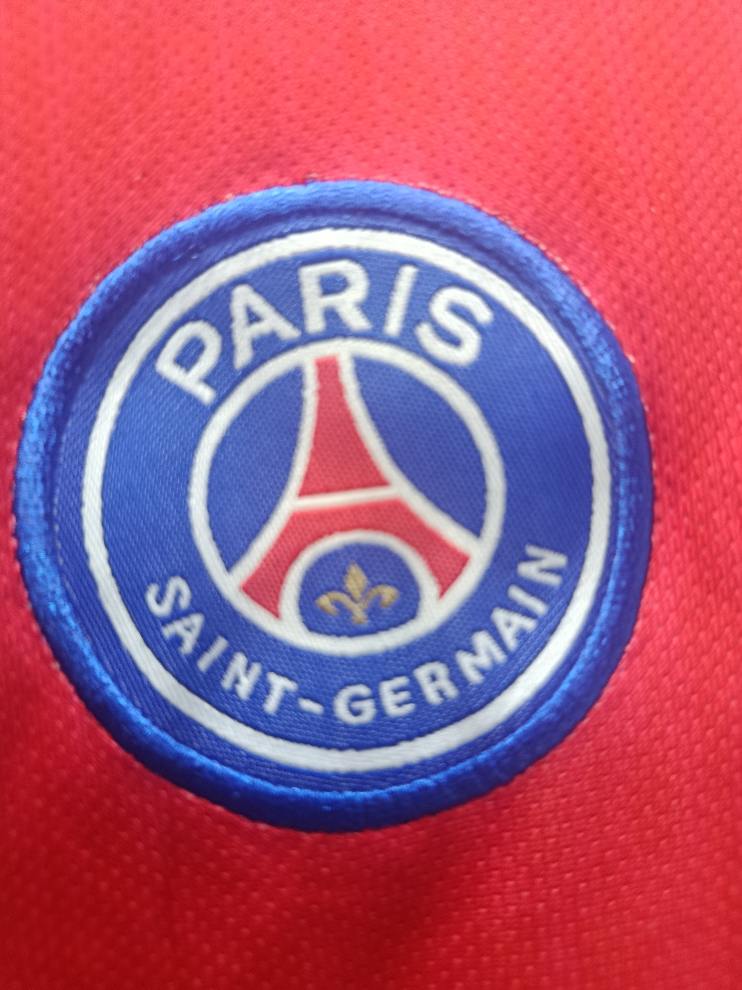 The image shows a close-up of a Paris Saint-Germain football club crest on a red garment. The crest is circular with a blue outer ring and a white inner ring. Inside the inner ring, the word "PARIS" is displayed in white capital letters at the top. Below "PARIS," a red Eiffel Tower silhouette is visible. The bottom portion of the crest contains the words "SAINT-GERMAIN" in white capital letters, curved along the bottom. A gold fleur-de-lis symbol is located beneath the Eiffel Tower. The red fabric of the garment is textured with small, raised dots. There is no indication of the city of Sertolovo, Russia, in the image.