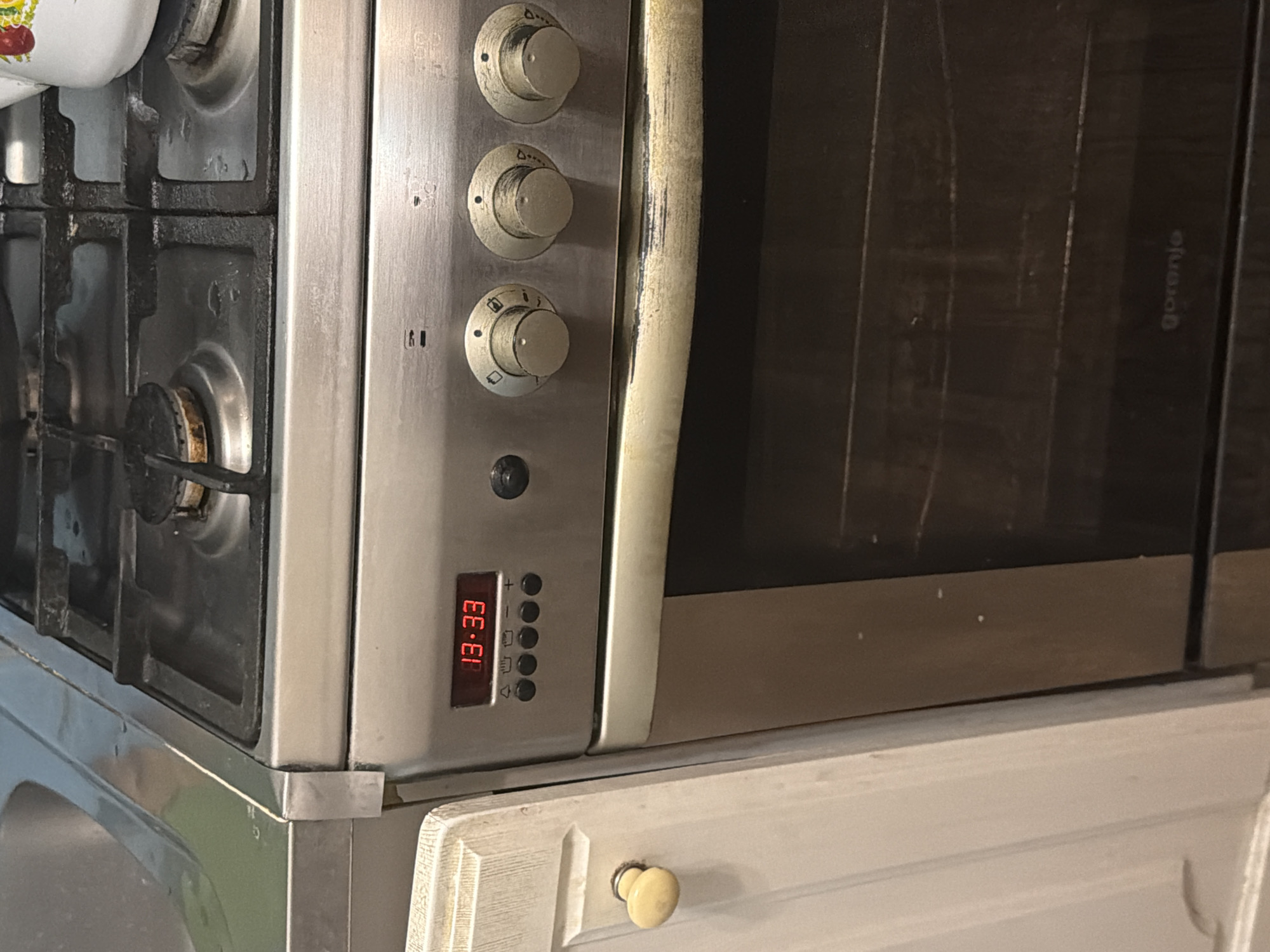 A close-up, eye-level shot shows the control panel and stovetop of a stainless steel gas oven. The digital display on the oven reads "13:33". Below the display are four circular buttons and a plus and minus button. To the right of the buttons are three rotary knobs, each with markings indicating different settings. The stovetop, which is black and appears to be made of cast iron, has four burners, one of which has a flame visible. The front of the oven has a silver handle that appears to be discolored with a yellowish-brown grime. To the left of the oven is a white cabinet door with a round, off-white knob. A stainless steel countertop with a curved edge is visible in the upper left corner of the image, with a portion of a sink peeking out. The overall environment appears to be a kitchen.