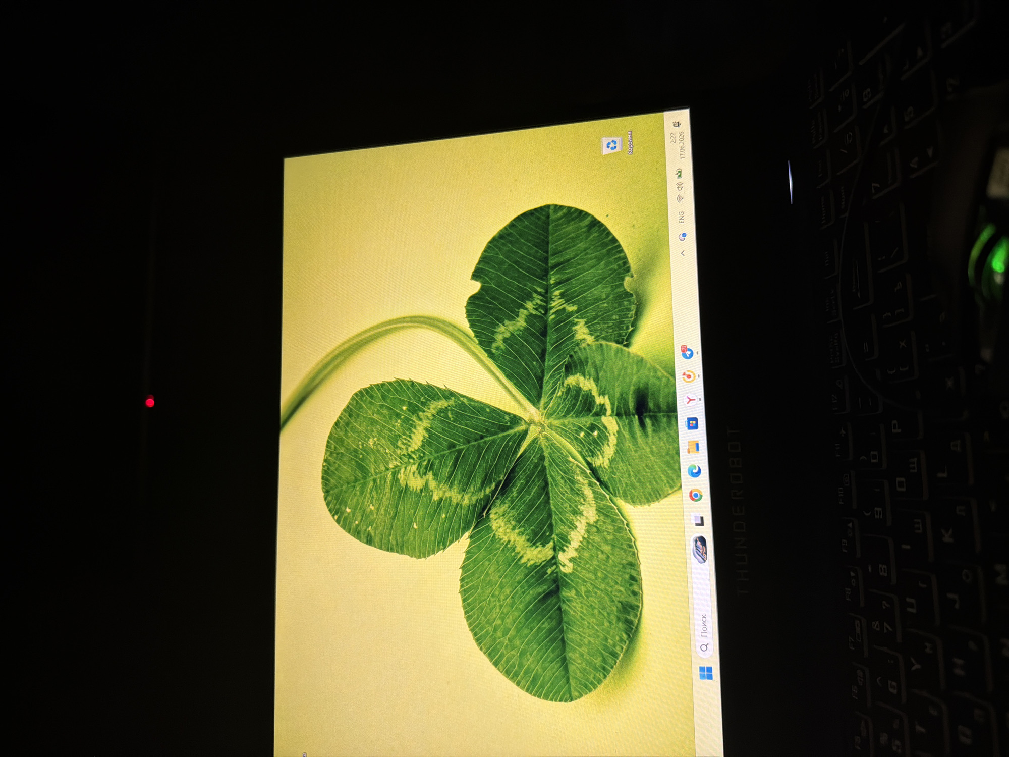 The image shows a close-up view of a laptop screen displaying a four-leaf clover wallpaper. The screen is illuminated against a dark background, suggesting the room is dimly lit. The laptop itself is a THUNDEROBOT brand, visible on the chassis below the screen. The keyboard is also visible, with illuminated keys. The bottom right corner of the screen shows the Windows taskbar with icons for search, language (ENG), audio, and the date and time: 2:22, 17.06.2026. A red LED is visible in the upper part of the dark background, possibly indicating a power or status light on the laptop or surrounding device. The location is identified as Sirius, Russia.