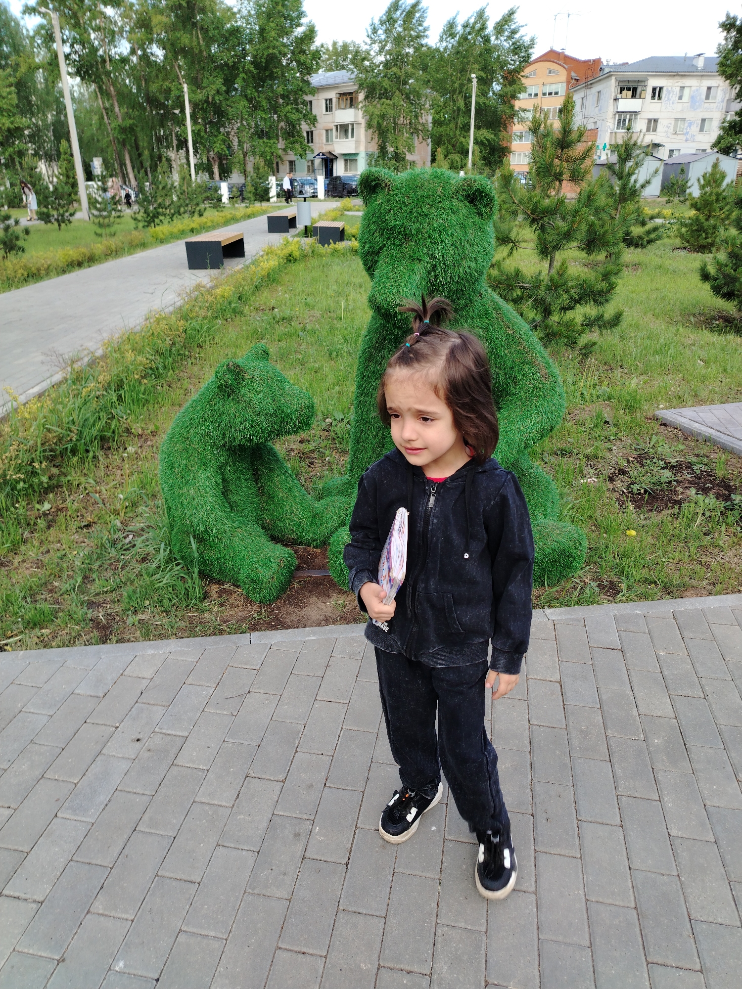 The image shows a young girl standing on a brick-paved area. She is wearing a dark-colored tracksuit and black and white sneakers, and she holds a notebook in her left hand. Behind her are two green bear-shaped topiary sculptures on a grassy lawn, with other trees and buildings visible in the background. The location appears to be a park or public space within the city of Chaikovsky, Russia.