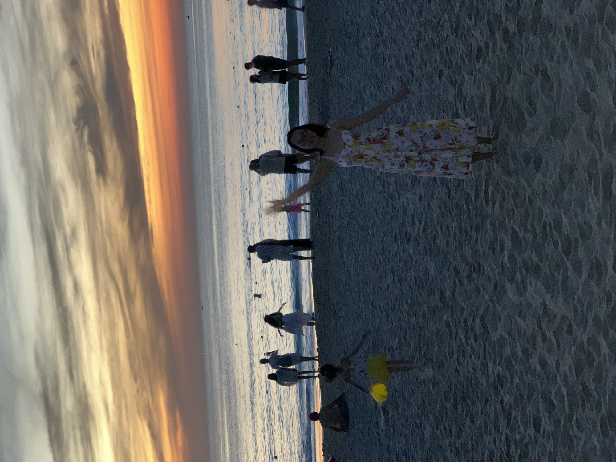 The image captures a beach scene at sunset in Da Nang, Vietnam. The sky is ablaze with hues of orange, yellow, and grey from the setting sun, with wispy clouds stretching across the horizon. The calm sea reflects the colors of the sky. On the sandy beach, several people are visible. In the foreground, a woman in a floral dress stands with her arms outstretched, appearing to wave or pose. Further back, a young girl in a yellow skirt is holding a yellow bucket. Other individuals and groups are scattered across the beach and near the water's edge, some walking, some standing, and one person is sitting. The overall atmosphere suggests a relaxed evening by the coast.