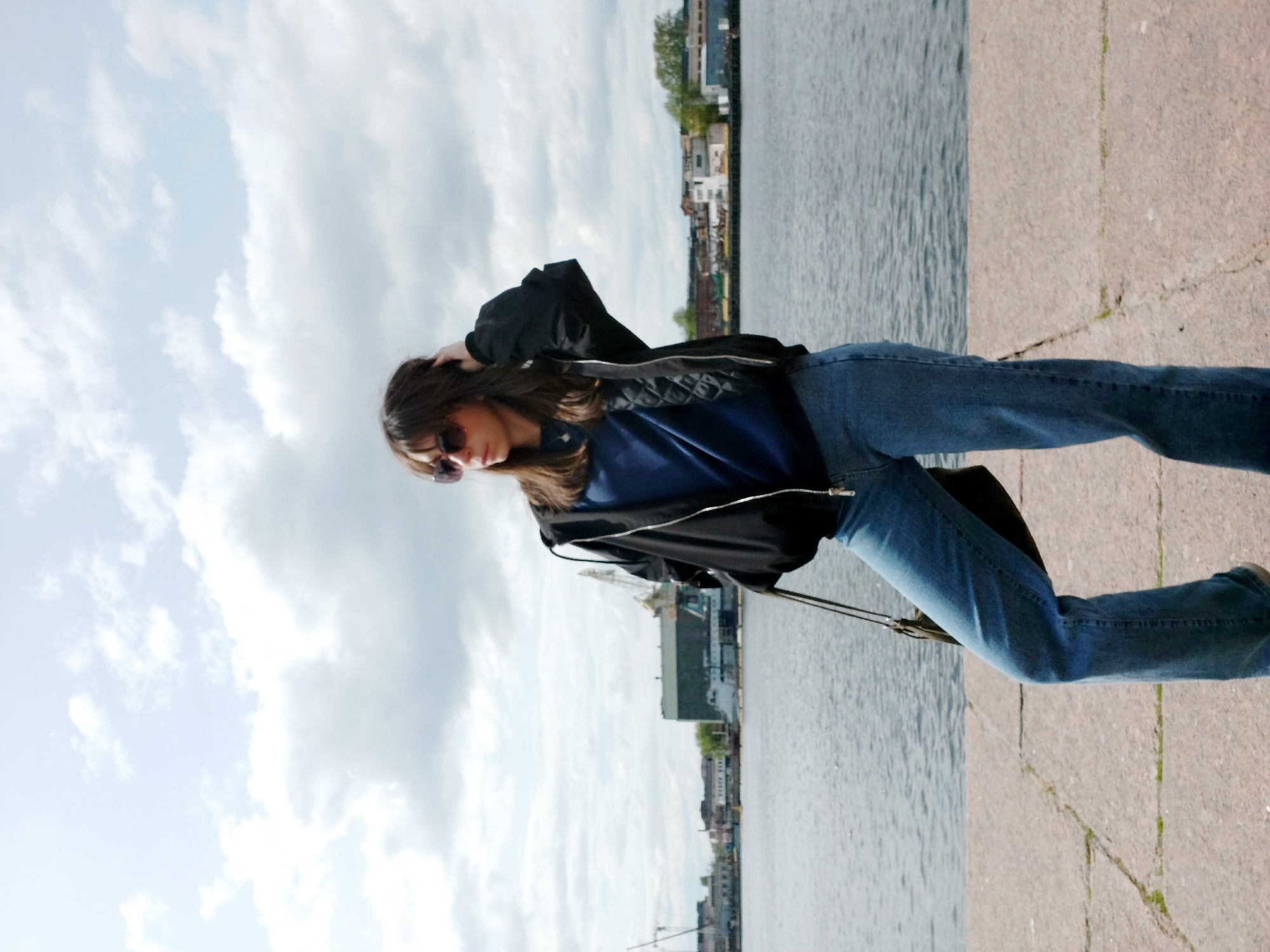 A full body shot of a woman standing on a paved surface next to a body of water. The woman has brown hair and is wearing sunglasses, a blue shirt, a black jacket, and blue jeans. She has one hand on her head. The body of water has buildings and some trees visible across the water. The sky is cloudy. The location is in an unknown city.