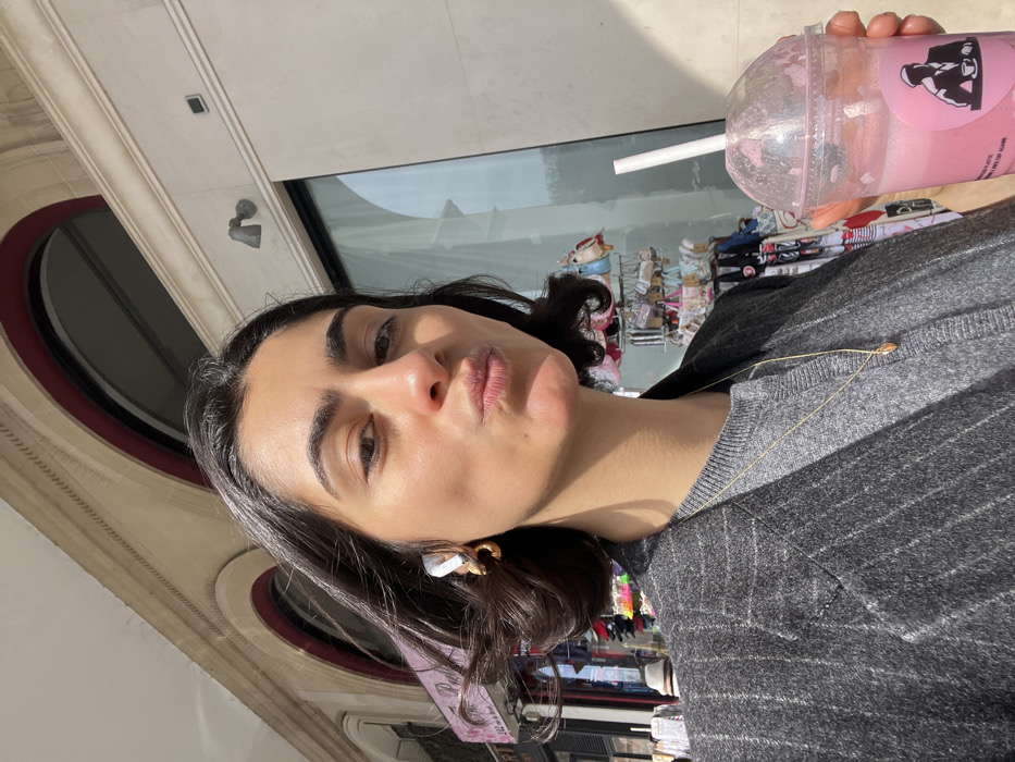 A female individual is captured in a close-up, upward-angle selfie on a sunny day in Paris, France. The subject has dark, medium-length wavy hair, dark eyes, and a light-medium skin tone, with her lips pursed in a pout. She wears a grey pinstriped blazer over a grey knit sweater and a delicate gold chain necklace with a small pendant. A white wireless earbud is visible in her left ear, accompanied by a gold-colored hoop earring. In her right hand, she holds a clear plastic cup containing a pink beverage with a white straw. The cup features a circular black and white sticker depicting a silhouetted man in a hat holding a coffee cup, with the text "MAN CLUB" partially visible.
The background on the left reveals an outdoor street scene with various merchandise displayed on racks, including small bags and hats, suggesting a market or souvenir vendors. A partial sign with "242" and "Rue de..." is visible, identifying the location as Rue de Rivoli, Paris, France, specifically around number 242. On the right, a modern building facade with large reflective glass windows and light-colored stone or concrete architecture is visible, featuring an arched window opening above. A small fixture, possibly a security camera or light, is mounted on the wall above the window. The overall environment suggests a bustling urban street.