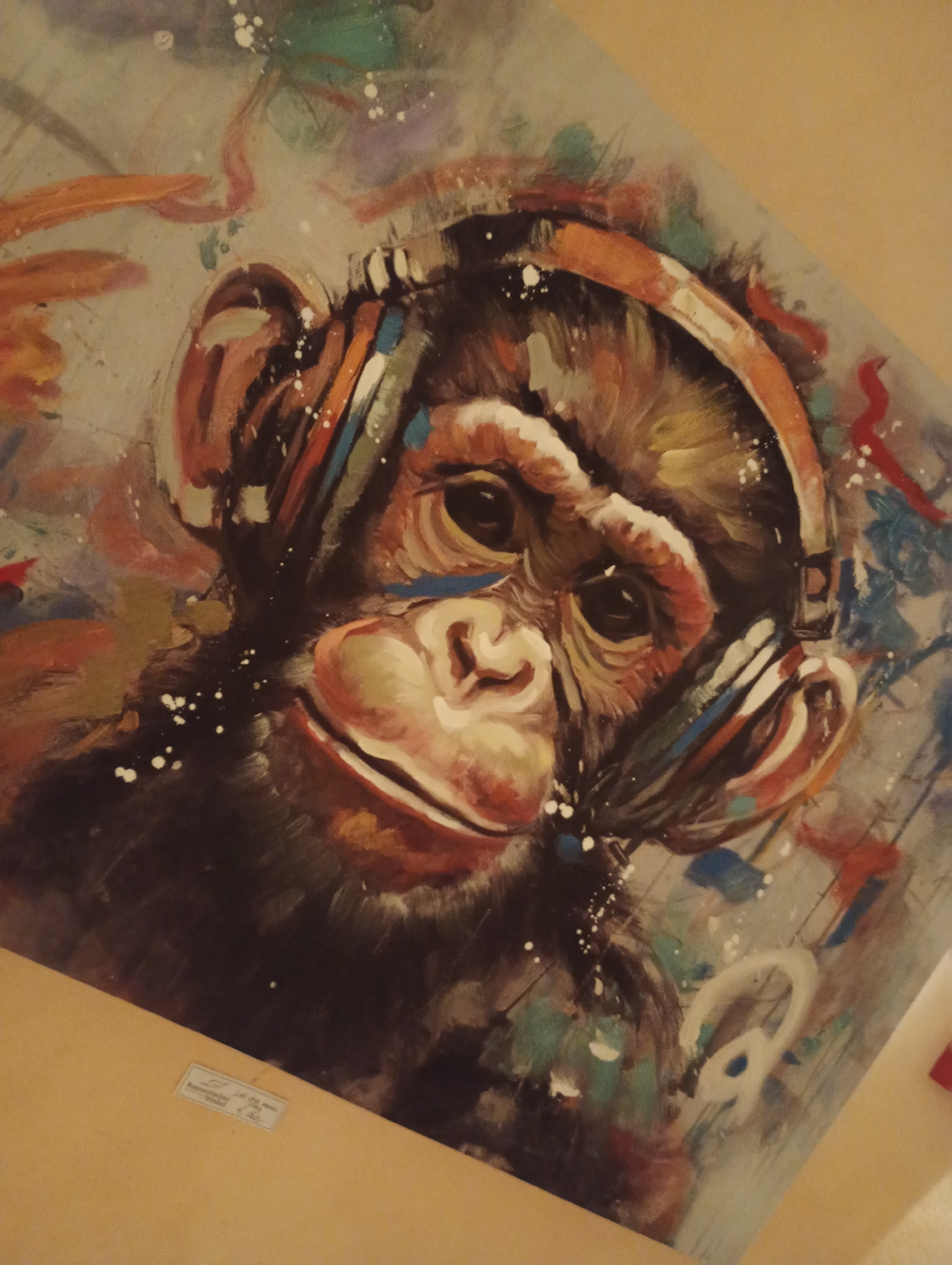 A framed painting of a chimpanzee wearing headphones is displayed on a wall. The painting is abstract and colorful, with visible brushstrokes. The chimpanzee's face is the central focus, with headphones covering its ears. Splashes of paint in various colors, including white, orange, blue, and green, surround the chimpanzee. A small, light-colored label is affixed to the lower left corner of the frame, but the text is illegible. The background appears to be a light beige color, suggesting the painting is hanging in an interior space. The image quality is slightly blurry, and the painting is tilted, indicating it might be a photograph taken at an angle. The overall scene depicts a piece of art in a domestic or gallery setting. The specific location within Bad Sachsa, Germany, cannot be determined from the provided image.