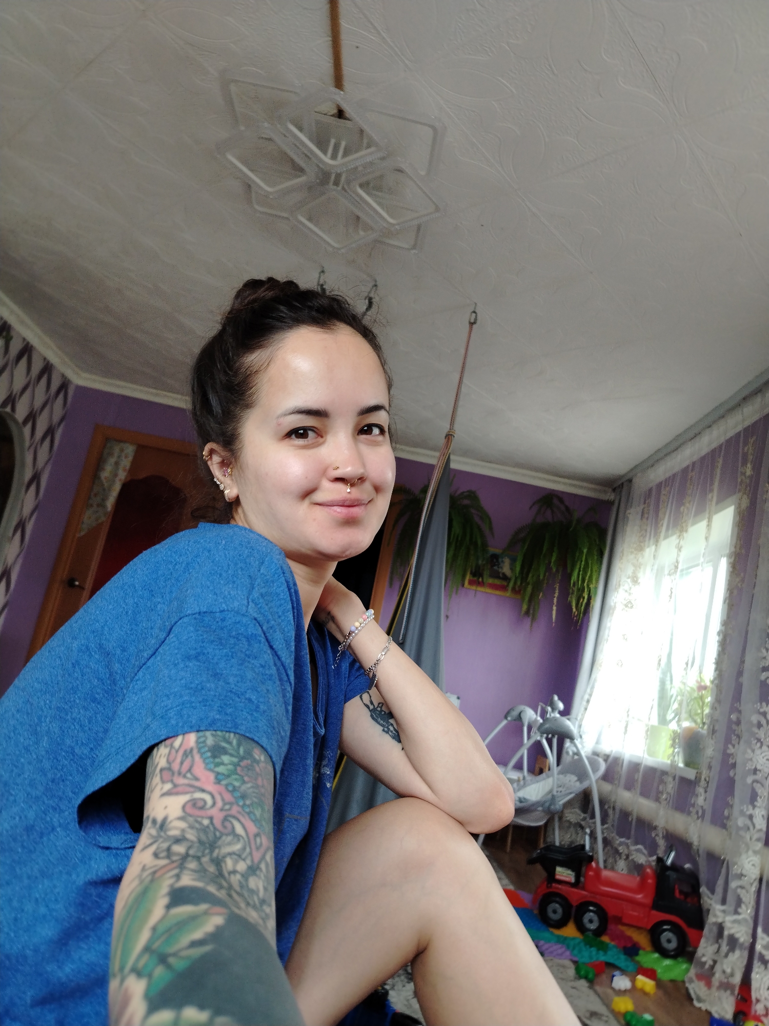 The image is a medium shot of a woman indoors. She has dark hair styled in a bun, fair skin, and facial piercings. She is wearing a blue t-shirt and has tattoos on her left arm. Her right arm is bent, and her hand is near her face. She is smiling. The background includes a textured white ceiling with a modern light fixture. The walls are painted purple, and a door is visible in the background. There is also a window with light-colored curtains. A baby swing and toy truck are visible on the floor. The location is Гожан, Russia.