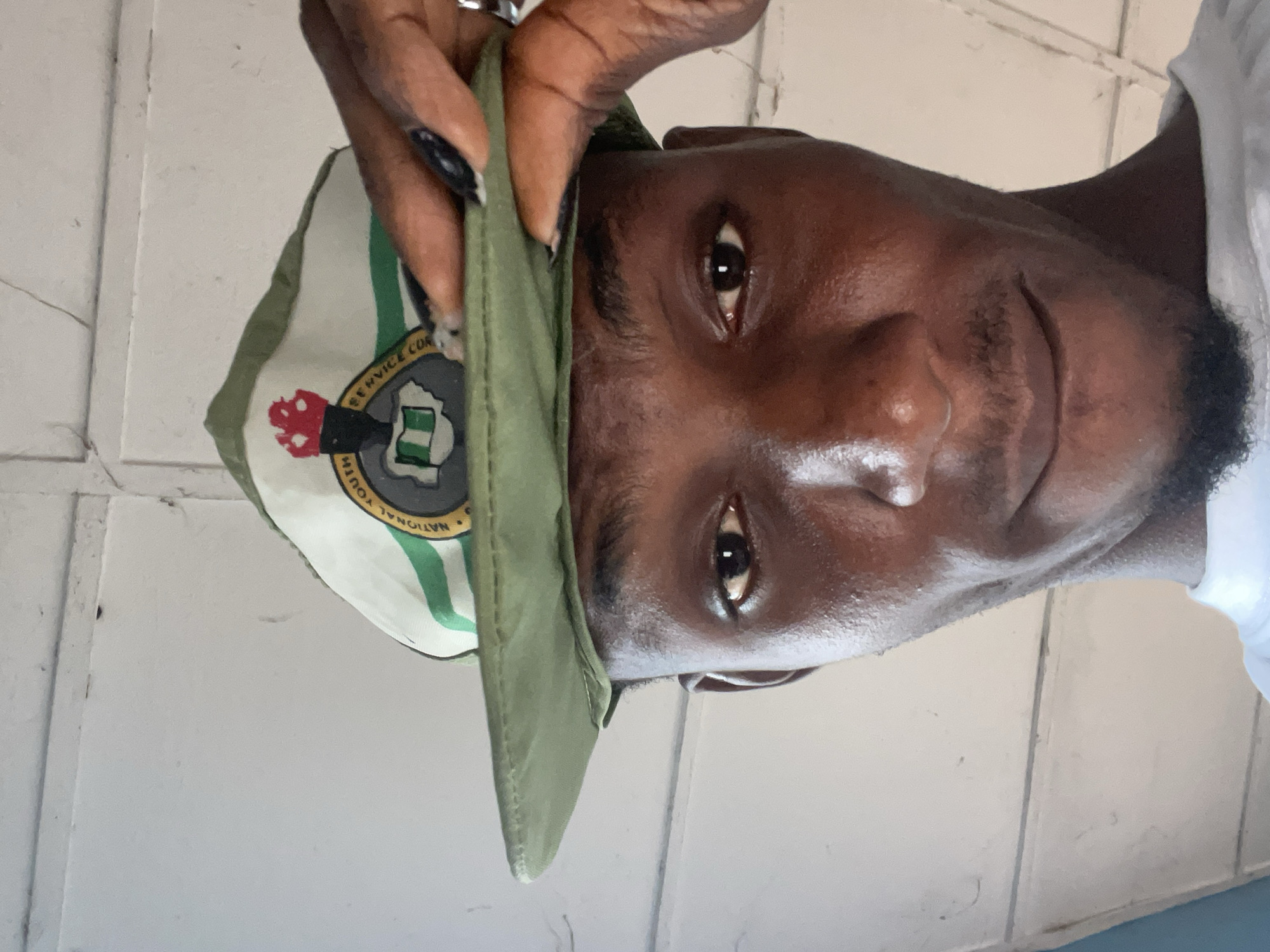 A low-angle, close-up selfie shows a young Black man wearing a green National Youth Service Corps (NYSC) cap. The cap has a white front panel with a circular logo featuring a torch, a map of Nigeria, and the text "NATIONAL YOUTH SERVICE CORPS". The man is holding the brim of the cap up with his right hand, revealing his forehead and eyes. His skin is dark and shiny, and he has short, dark hair and a small beard. He is looking directly at the camera with a slight smile. The background consists of a white, planked ceiling with visible lines and some minor imperfections. The overall lighting is bright, suggesting it's daytime. The image is taken in Ise-Ekiti, Nigeria.