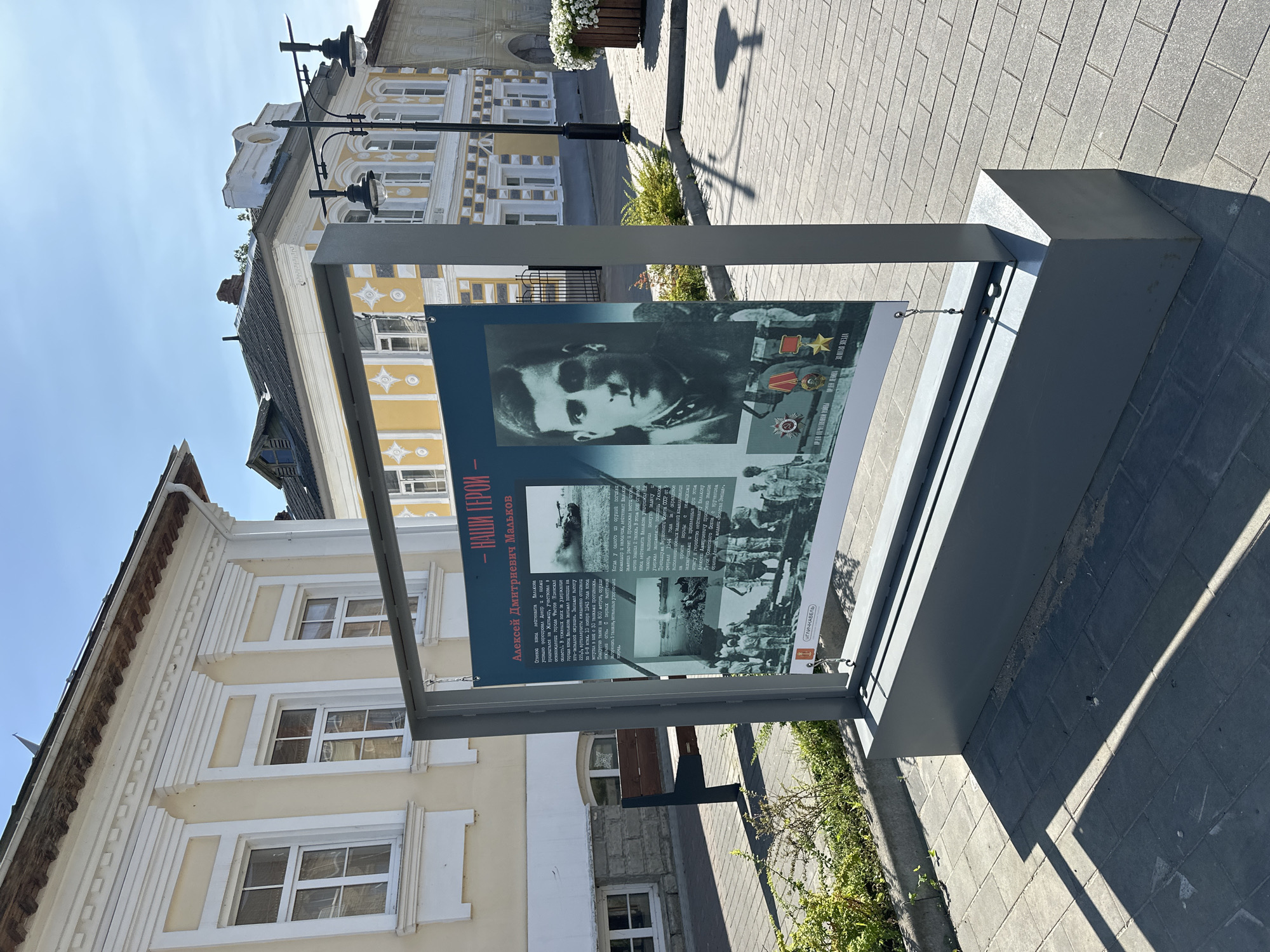 This is a medium shot of an outdoor informational display in the city of Nikul'skoe (Neverkovskii S/O), Russia. The display is housed in a grey metal frame and features a large banner with text and images. The banner appears to be a memorial or historical exhibit, with a portrait of a man in military uniform, along with other photographs of military scenes and medals. The text on the banner is in Russian, with the title "- НАШИ ГЕРОИ -" (Our Heroes) prominently displayed.
The display is situated on a paved walkway made of grey interlocking bricks. To the left of the display, there is a dark green park bench. Behind the display and bench, there is a building with a pale yellow facade and white decorative elements around the windows and roofline. Further in the background, other buildings with similar architectural styles can be seen, one of which is painted yellow with white ornamentation.
The scene is illuminated by bright sunlight, casting strong shadows on the ground. Green bushes are planted along the edge of the walkway. The sky is clear and blue. The overall impression is of a well-maintained public space, likely a town square or a pedestrian street.