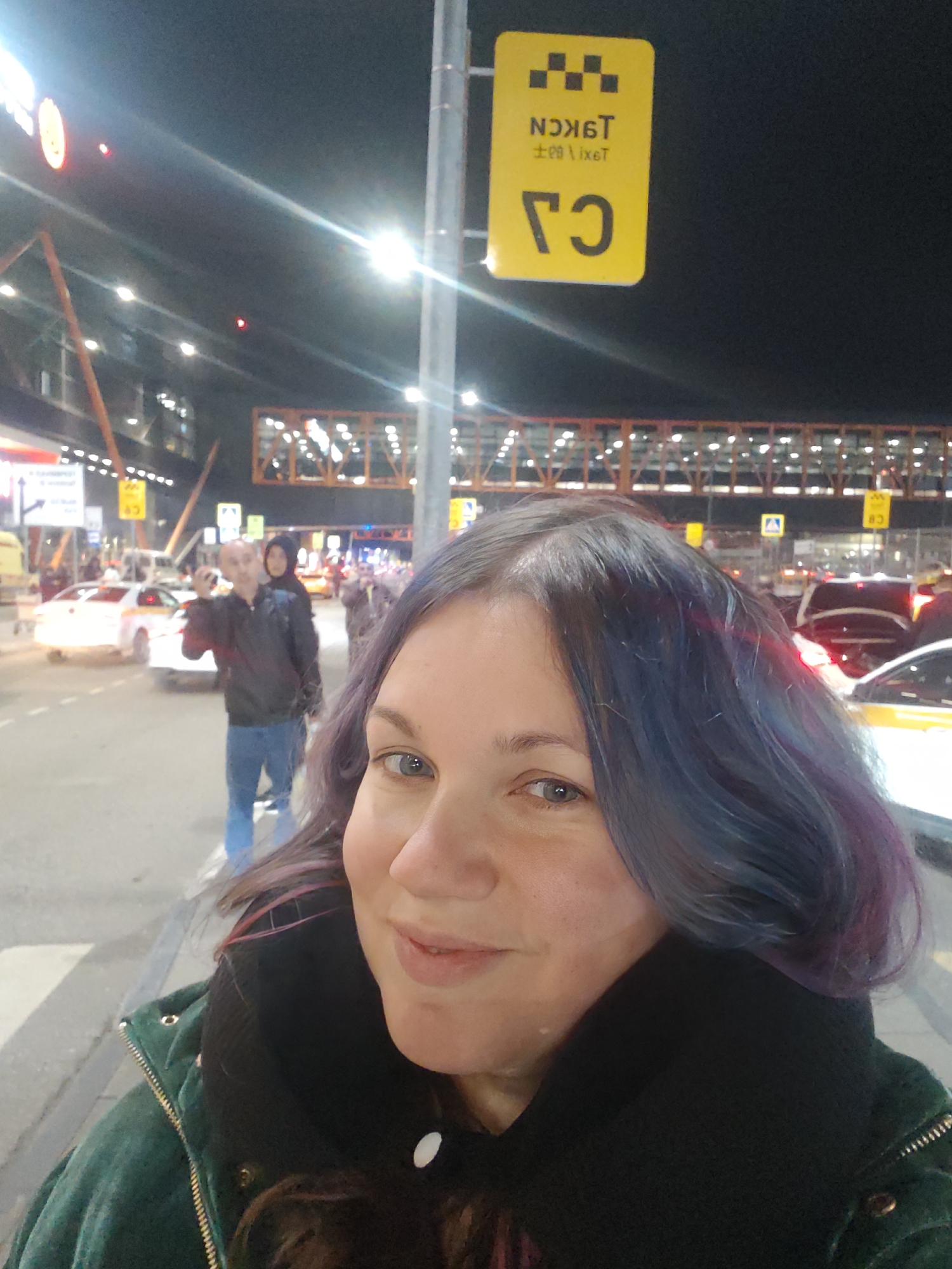 A selfie depicts a woman with dyed blue and purple hair, wearing a dark coat with a green jacket. The background shows a city scene at night with bright lights and various buildings. A yellow taxi sign with Cyrillic text is visible on a pole to the right. Behind her, there's a pedestrian walkway over a road with cars and people; one man is taking pictures. The scene is located in Khimki, Russia.