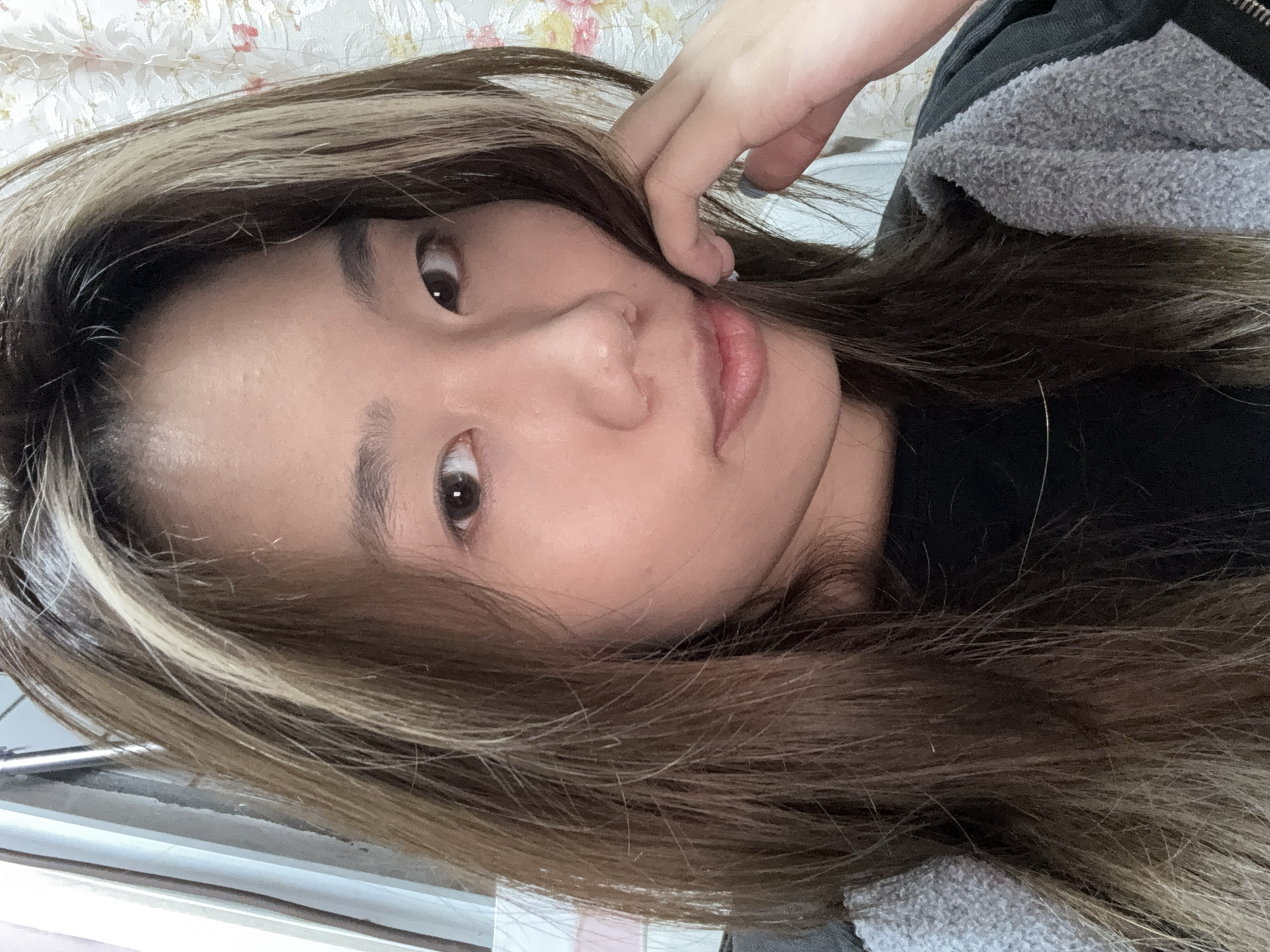 A close-up, slightly rotated, portrait of a woman with brown hair and blonde highlights. She is wearing a black shirt and a grey jacket. Her right hand is near her face. The background includes a floral pattern on fabric or wallpaper. No evidence in the image indicates a specific location in Shapingba District, China.