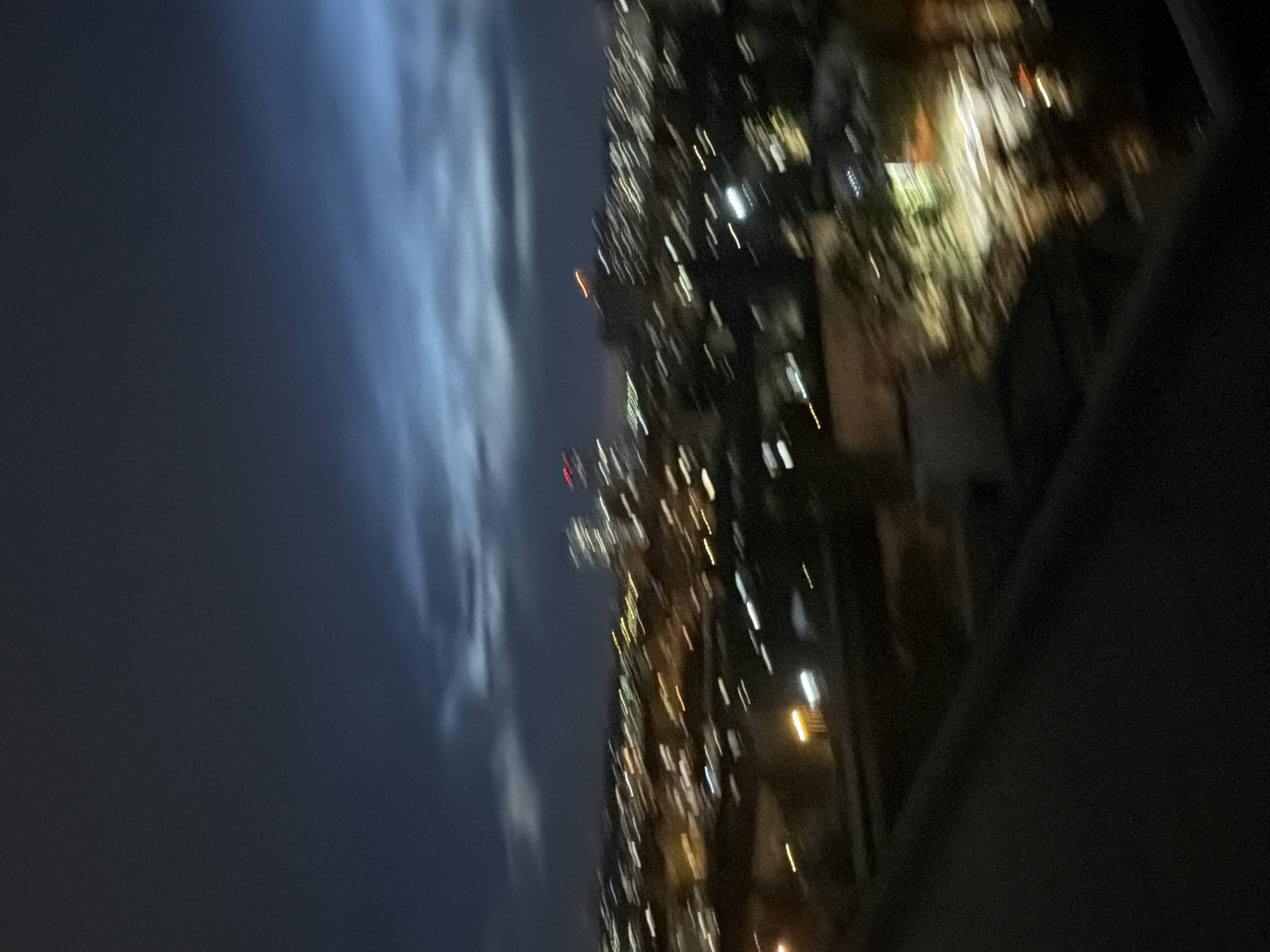 The image is a blurry, long-exposure photograph taken at night, overlooking a cityscape. The sky is dark blue with streaks of lighter blue and gray clouds, suggesting twilight or a stormy evening. Below the sky, the city lights are visible as elongated streaks of white, yellow, and orange, indicating motion or a long exposure. Some taller buildings with faint red and white lights can be discerned in the distance. The foreground is dominated by a dark, blurry surface, possibly a balcony railing or roof, with some faint lights visible in the background. The blurriness of the image makes it impossible to identify specific landmarks or details within the city, and therefore, it is impossible to determine the location as Kyiv, Ukraine. No people, actions, or interactions are discernible due to the lack of clarity. The environment appears to be an urban setting at night.