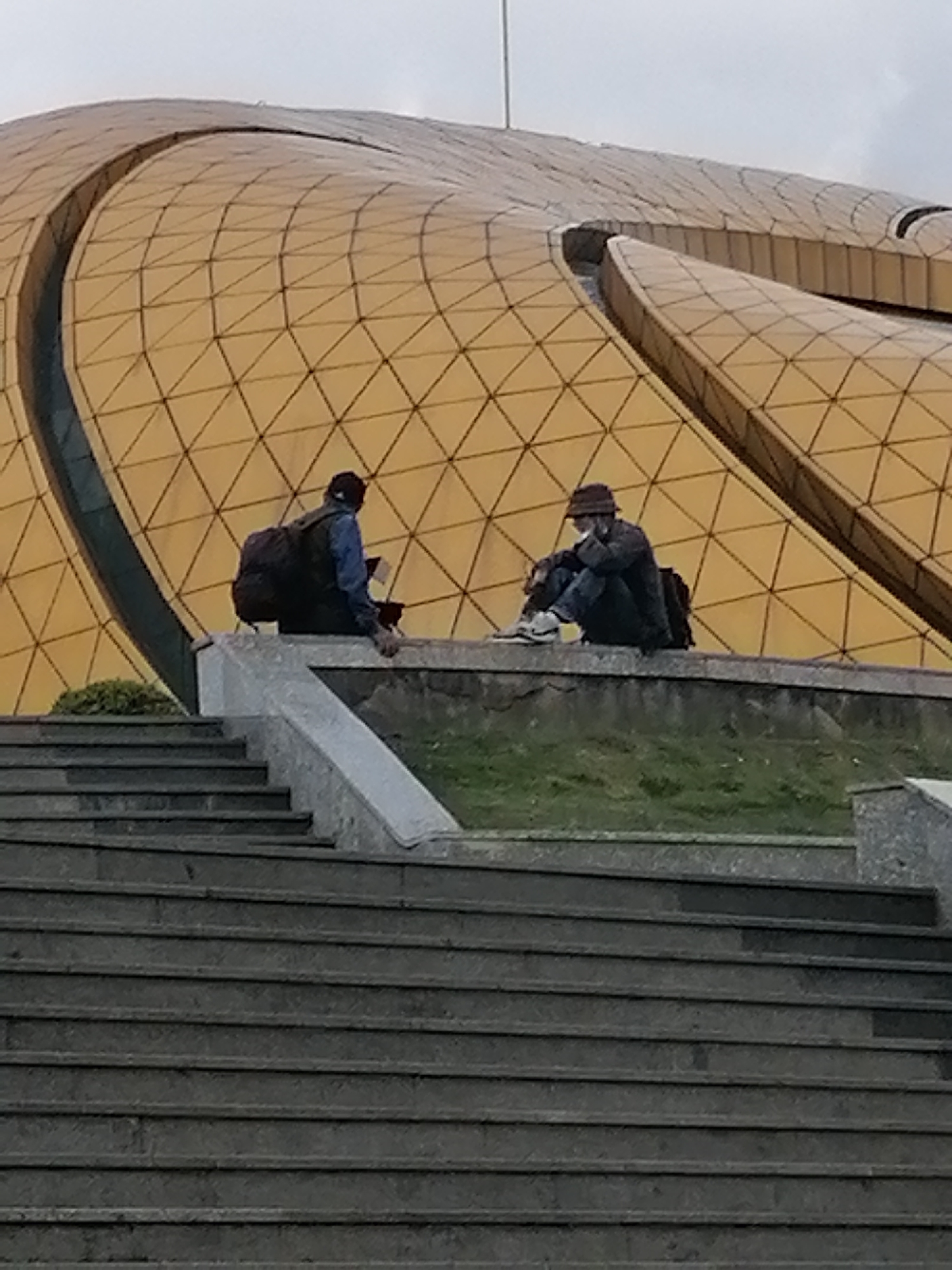 The image depicts two individuals sitting on a ledge in front of a large, golden, geodesic-dome-like building in Da Lat, Vietnam. The building's exterior is covered in a pattern of triangular panels. The two individuals are positioned on a concrete structure that appears to be a retaining wall or part of a tiered seating area, with stone stairs leading up from the foreground.
The person on the left is wearing a dark backpack and a blue jacket, and appears to be holding something in their hands. They are seated with their legs drawn up. The person on the right is wearing a hat and a dark jacket, and is also seated with their legs drawn up, leaning forward. Both individuals seem to be looking down or at something in front of them. The area around the ledge is covered in grass. The overall scene suggests a public outdoor space, likely a park or monument.